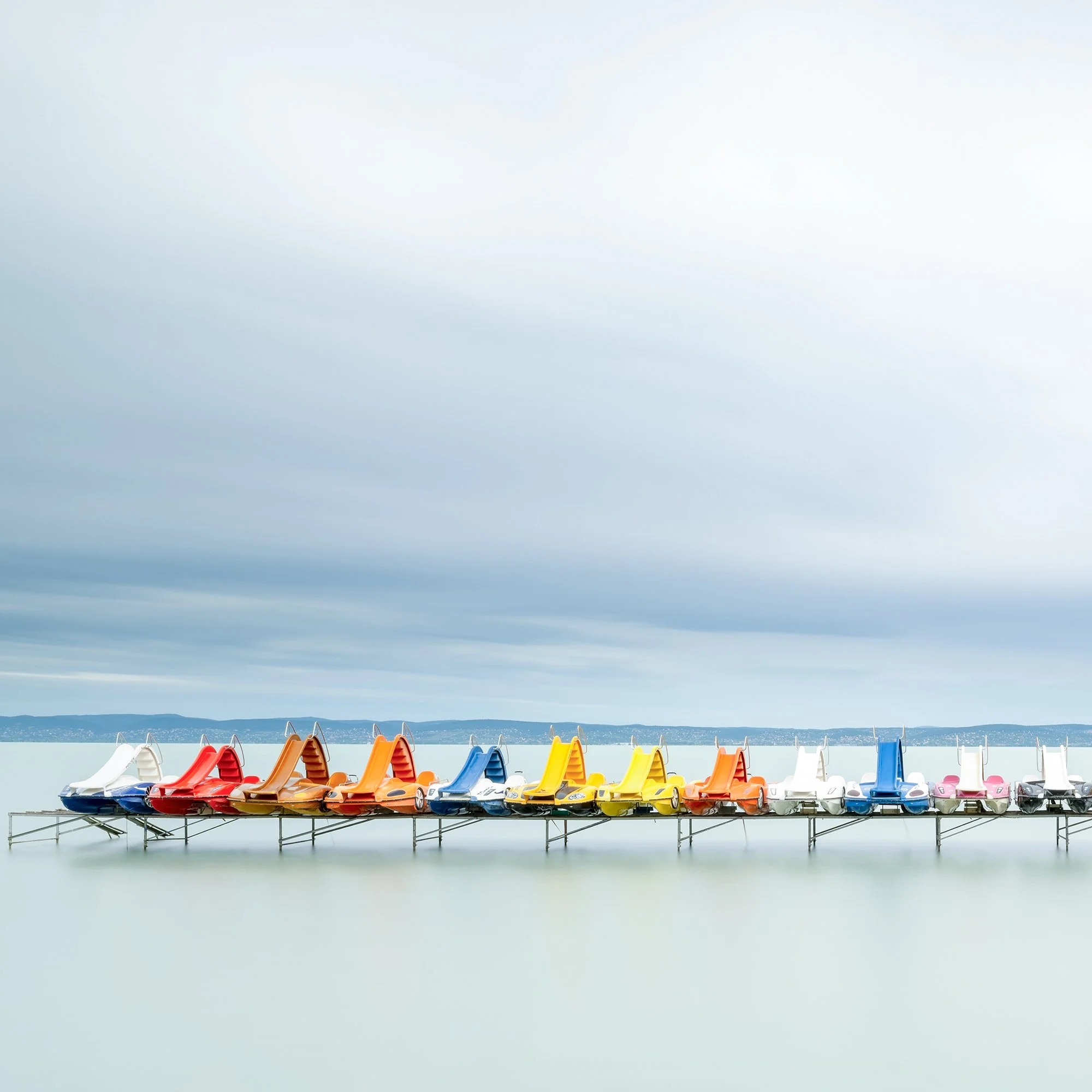 Gerald Berghammer - Color waterscape photography. Colorful pedal boats and a catamaran lined up on a dock on a calm body of water under a cloudy sky. Print detail 1