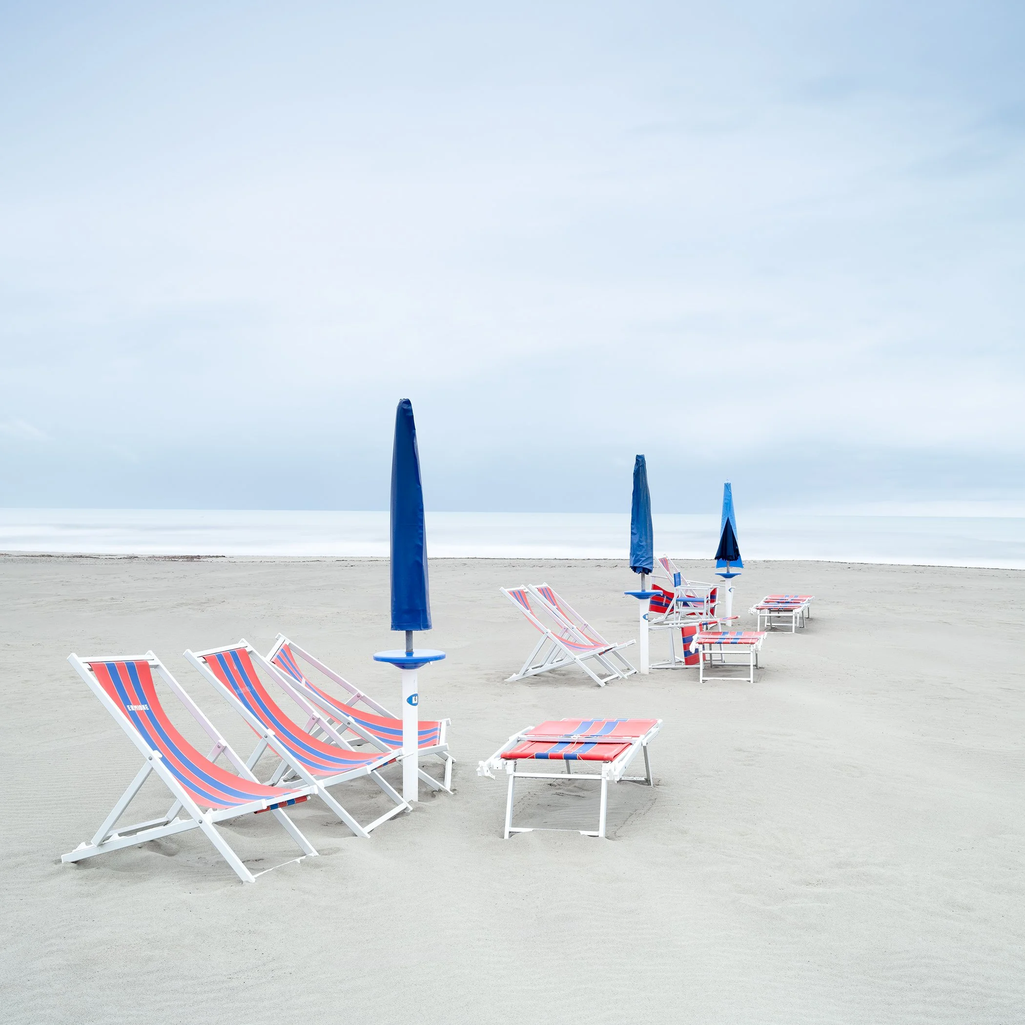 Minimalist colour photograph of empty sun loungers and closed parasols on a quiet beach in Italy. Photo by Gerald Berghammer.