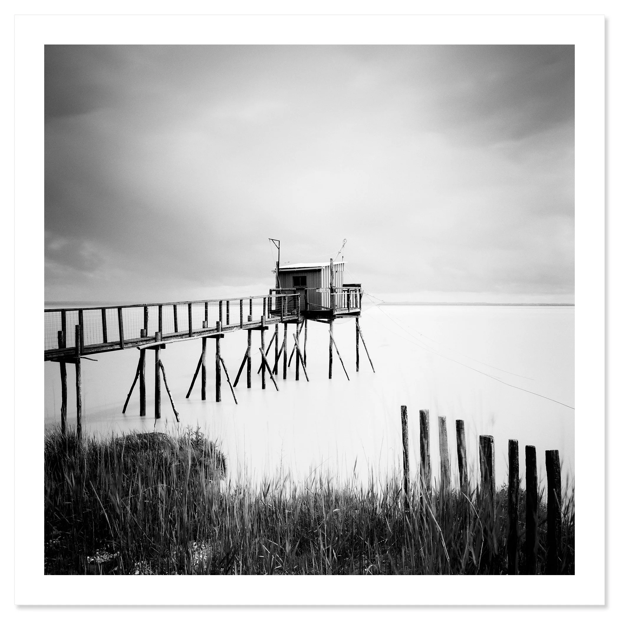 Gerald Berghammer - Black and white seascape photography. Small wooden hut on stilts connected by a long walkway over water with grass and a cloudy sky. Fine art print only