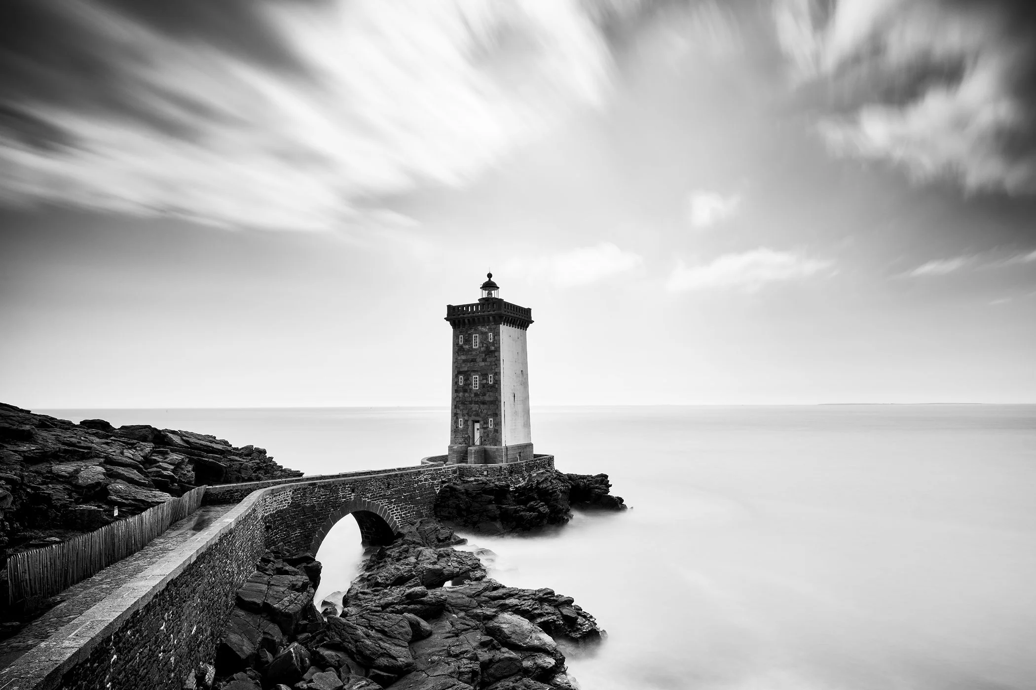 Black-and-white photo of the Kermorvan Lighthouse on a rocky coast, connected by a stone bridge