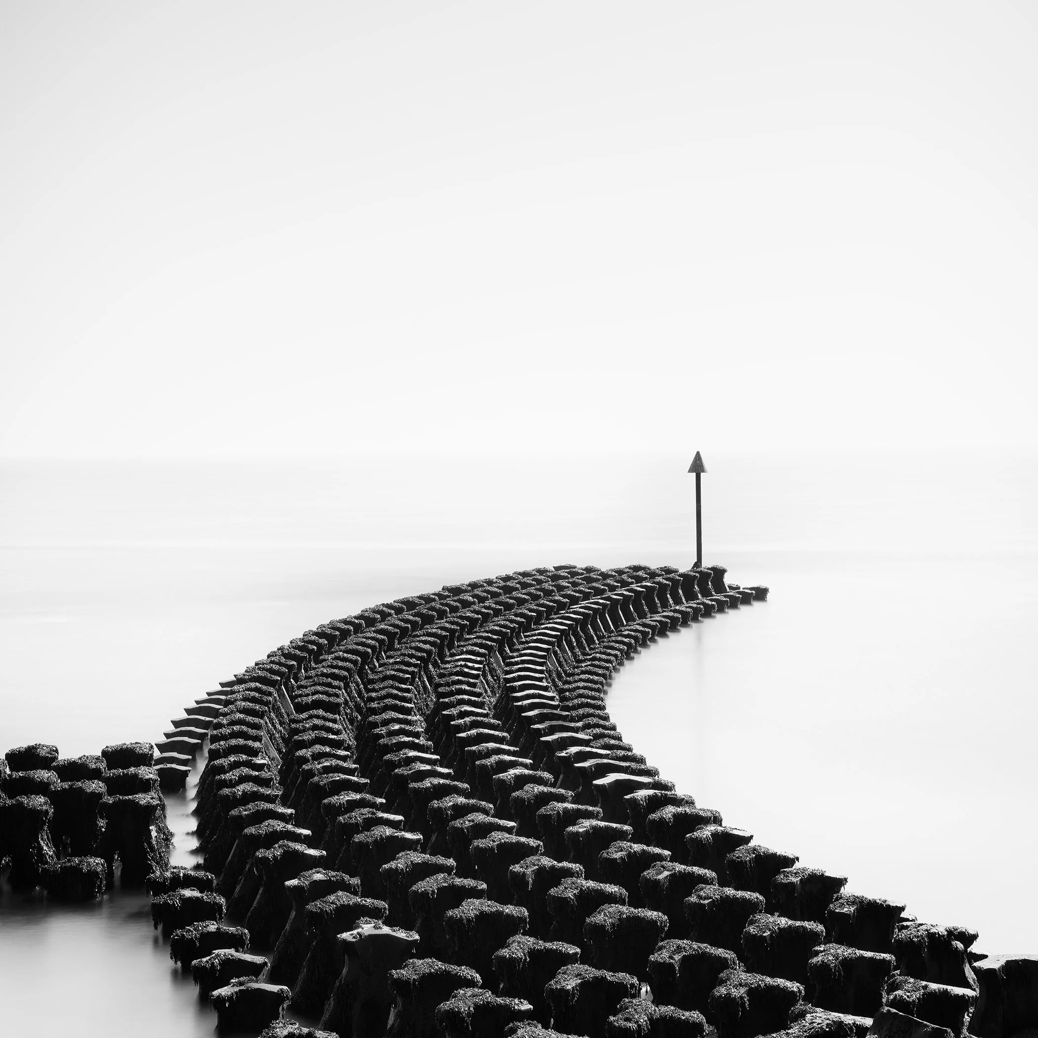 Curving wooden groynes extending into the calm North Sea at Cobbolds Point, England, photographed in black and white by Gerald Berghammer.