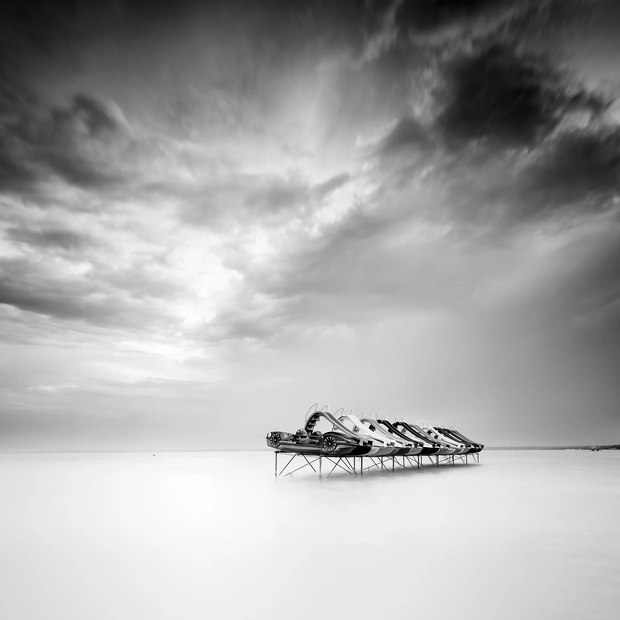 Pedal Boat Study 2, Lake Balaton, Hungary, monochrome long-exposure photograph of pedal boats on stilts in calm water under a cloudy sky.