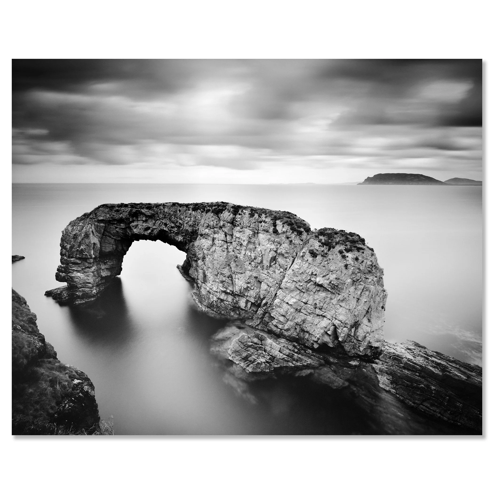 Gerald Berghammer - Black and white seascape photography. Natural rock sea arch formation in the ocean with an island on the horizon under a cloudy sky. Chromaluxe frameless