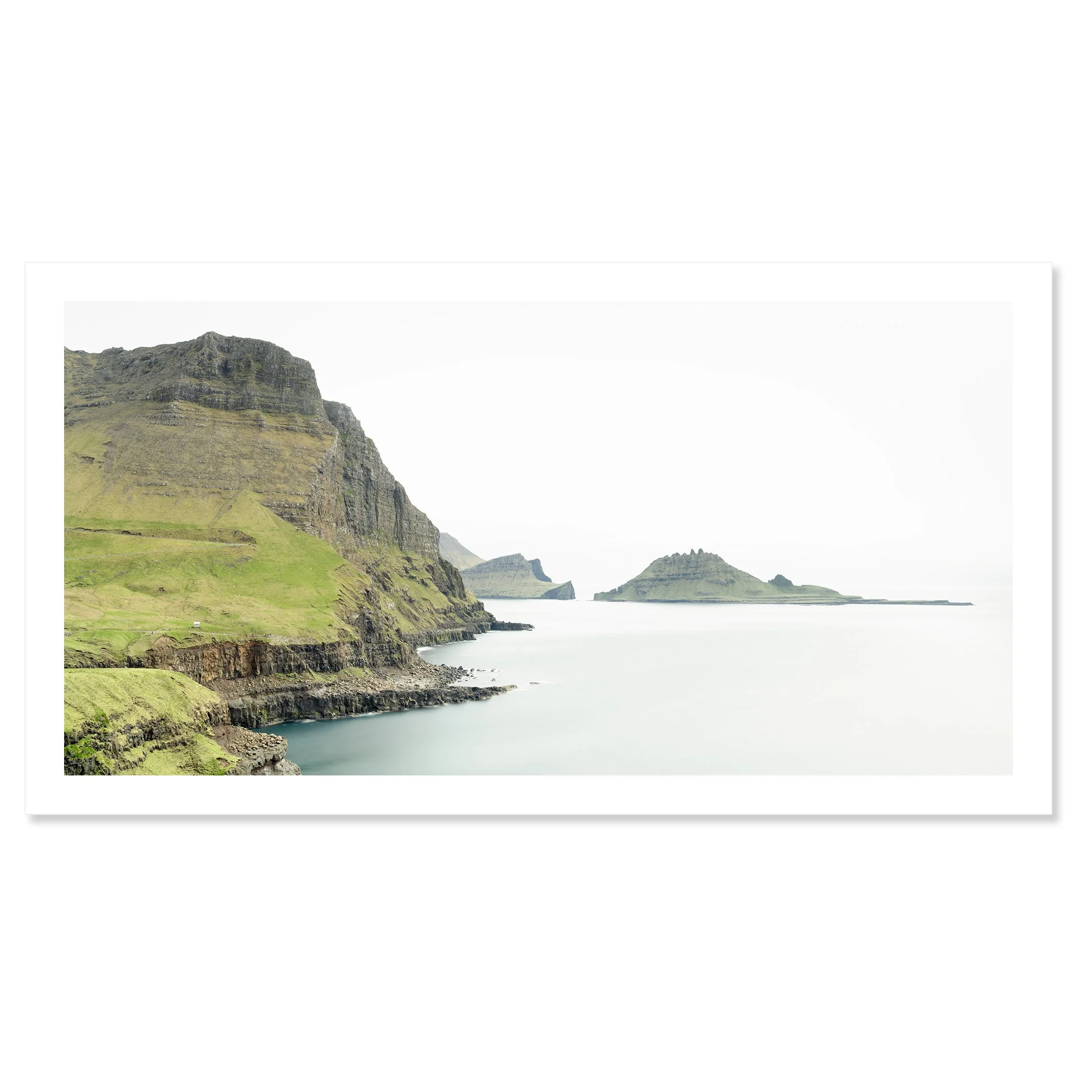 Gerald Berghammer - Color Photography. Cliffs and green hills along a body of water, with distant islands or landmasses in the background under a cloudy sky. Fine art print only