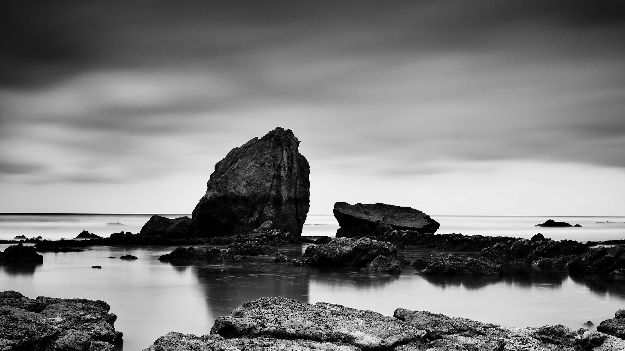 Black and white long-exposure seascape with large coastal rocks reflected in calm water under a cloudy sky