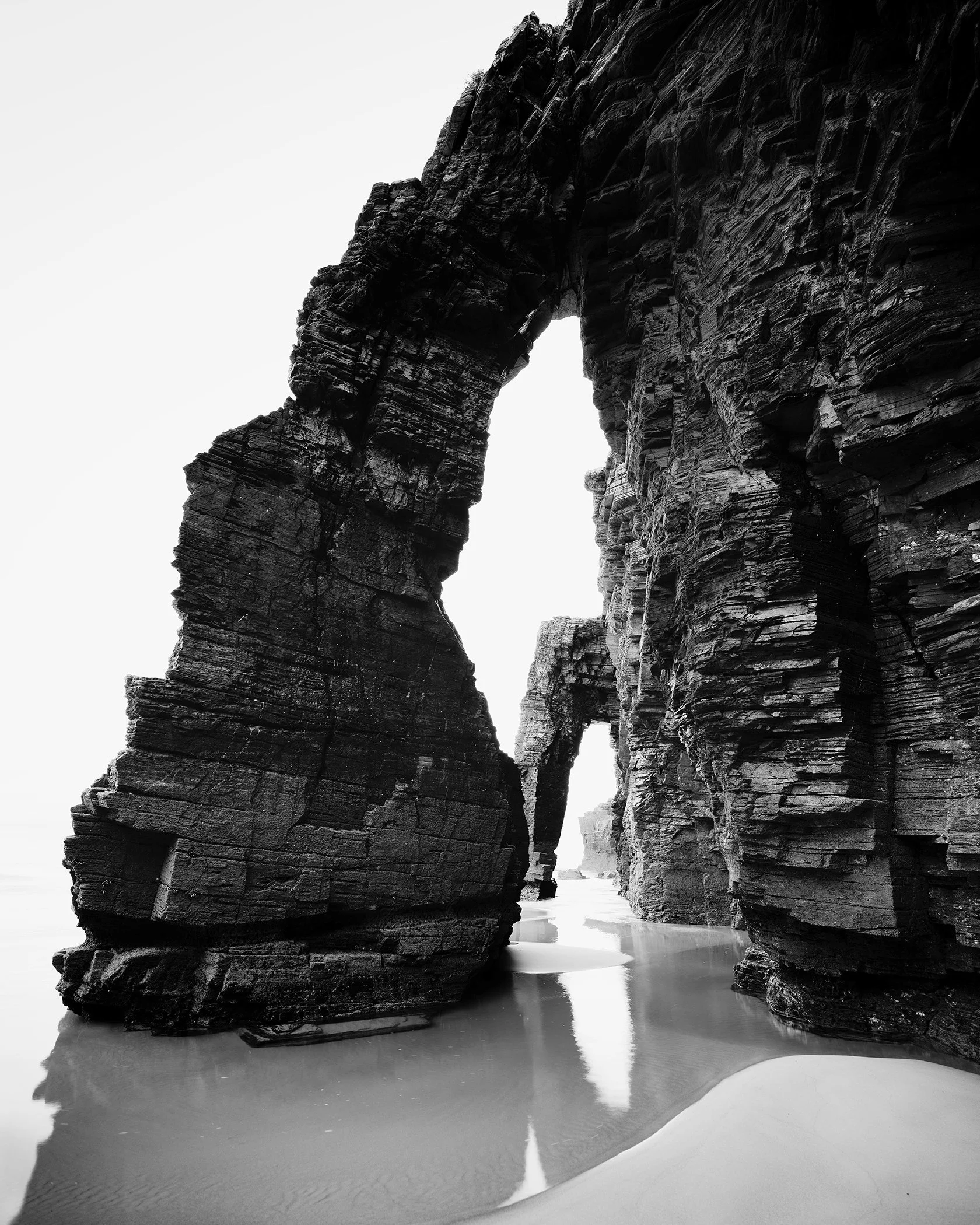 Long exposure black and white beach seascape showing rock arch formations, wet sand reflections and soft surf.