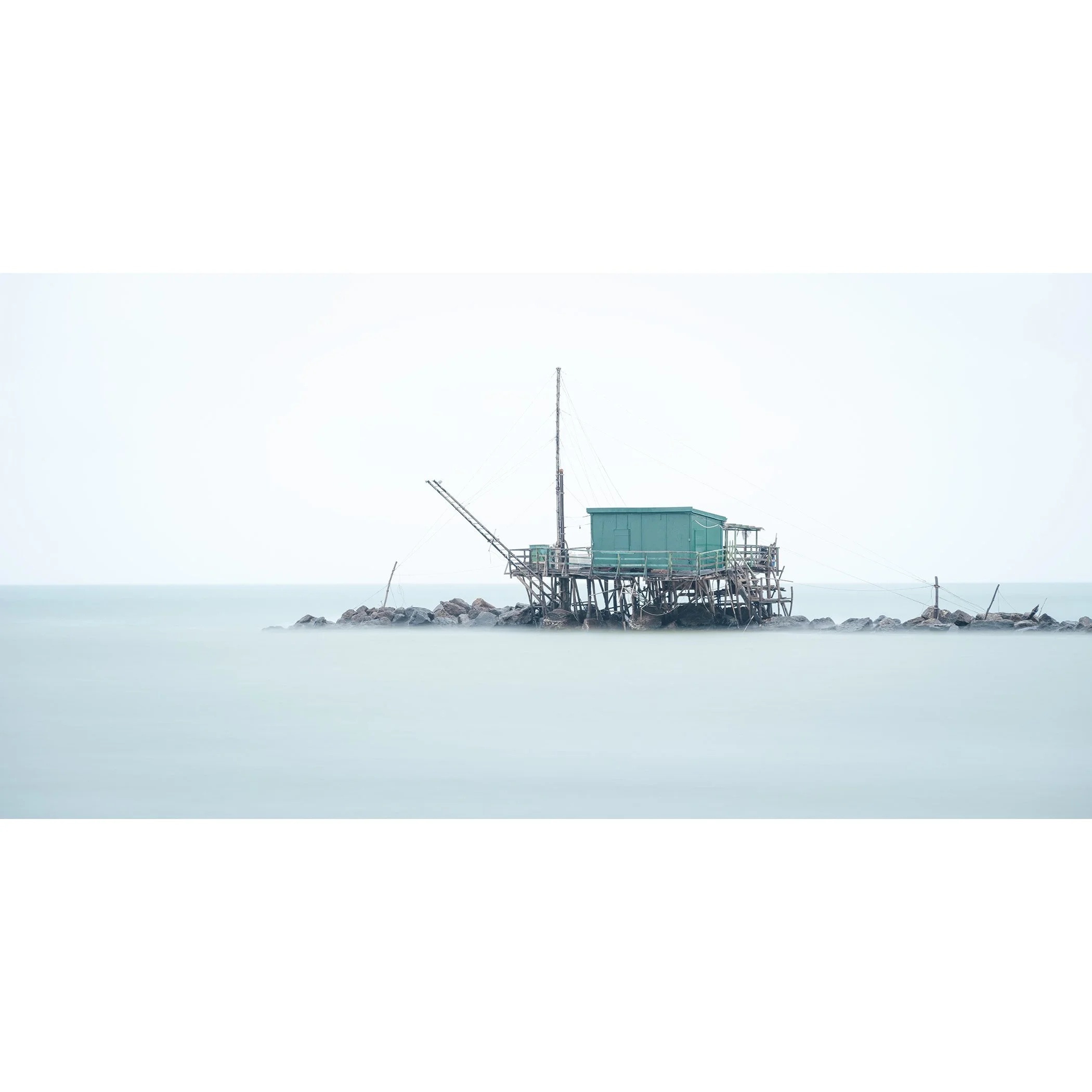 © 2024 Gerald Berghammer - Color minimalist photography. A small house built on stilts over water on rocks, with a fishing pole, in a minimalist, misty seascape.