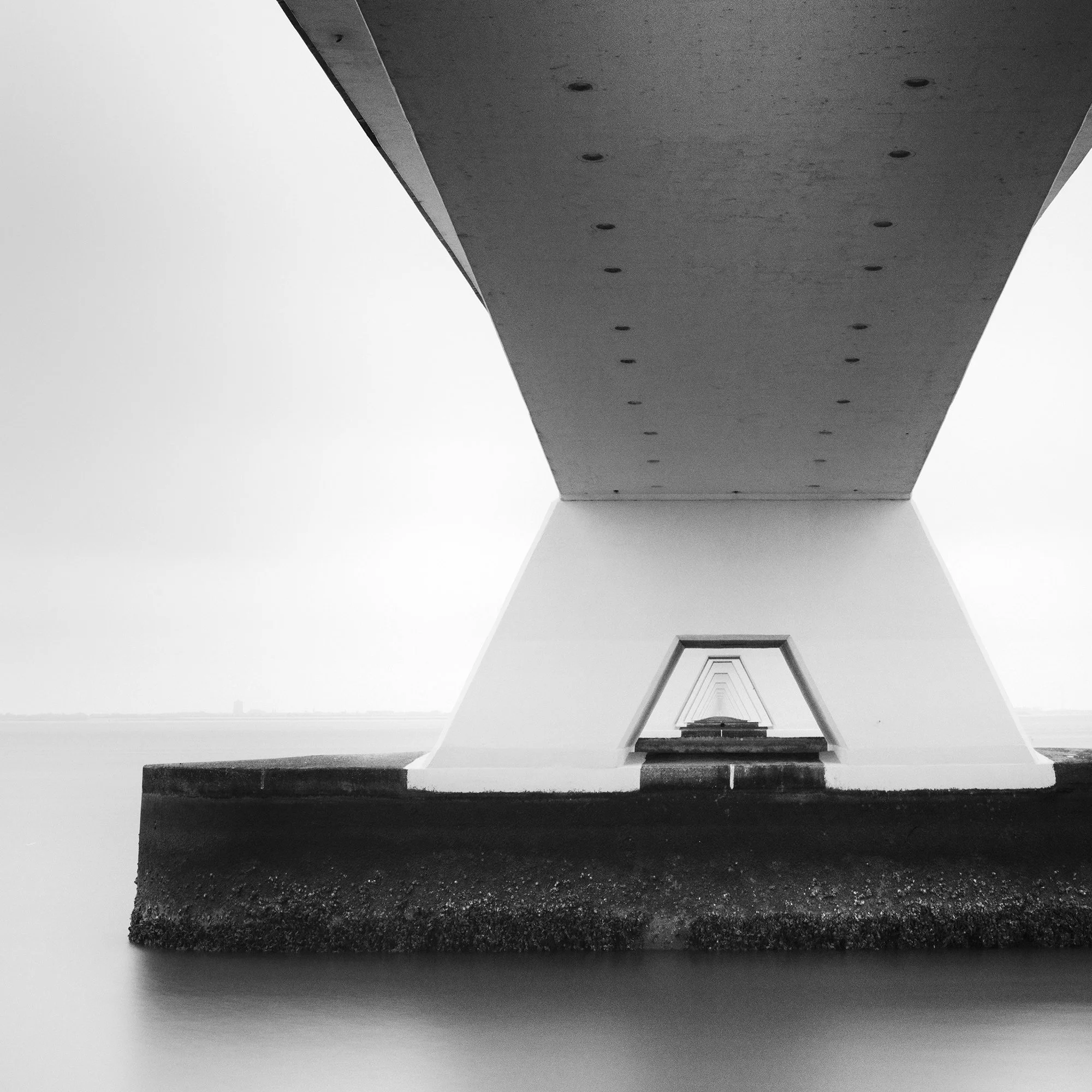 Gerald Berghammer - Black and white cityscape photo. Underneath view of a bridge with large support pillars, showing the underside and the water below. Print detail 1