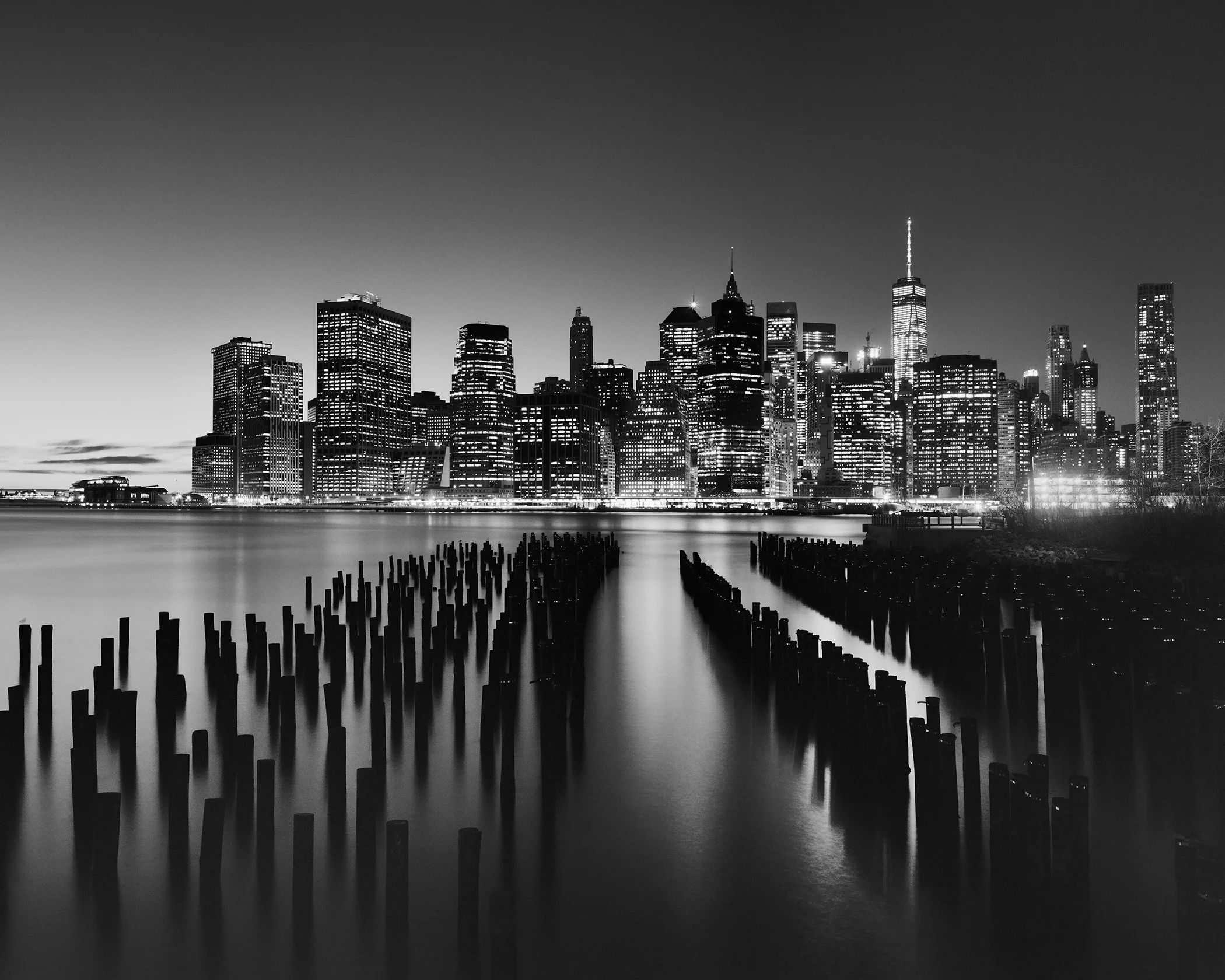 Manhattan Skyline Study 1 by Gerald Berghammer, black-and-white long-exposure photograph of Lower Manhattan, One World Trade Center and wooden pilings in the East River, New York City, USA.