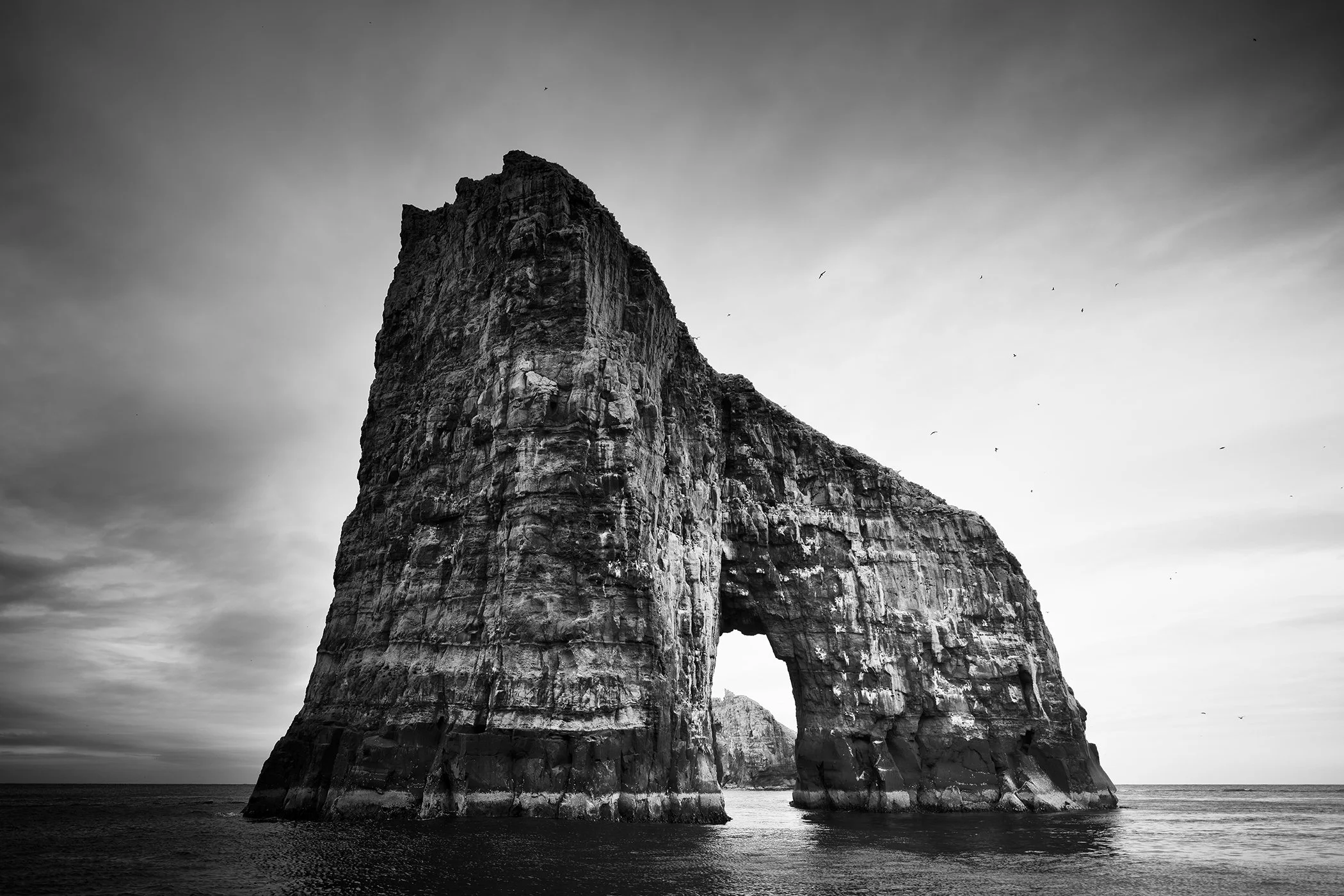 Black and white coastal landscape showing a towering sea stack with a natural rock arch