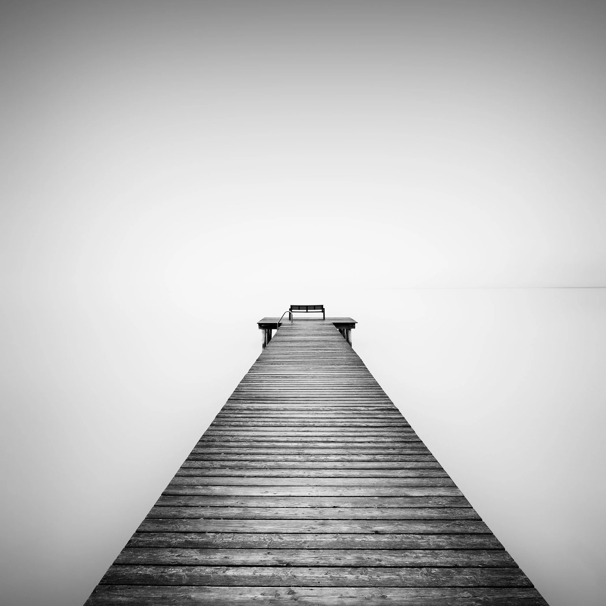 Minimalist black-and-white waterscape of a wooden jetty reaching into a calm misty lake in Austria, by Gerald Berghammer.