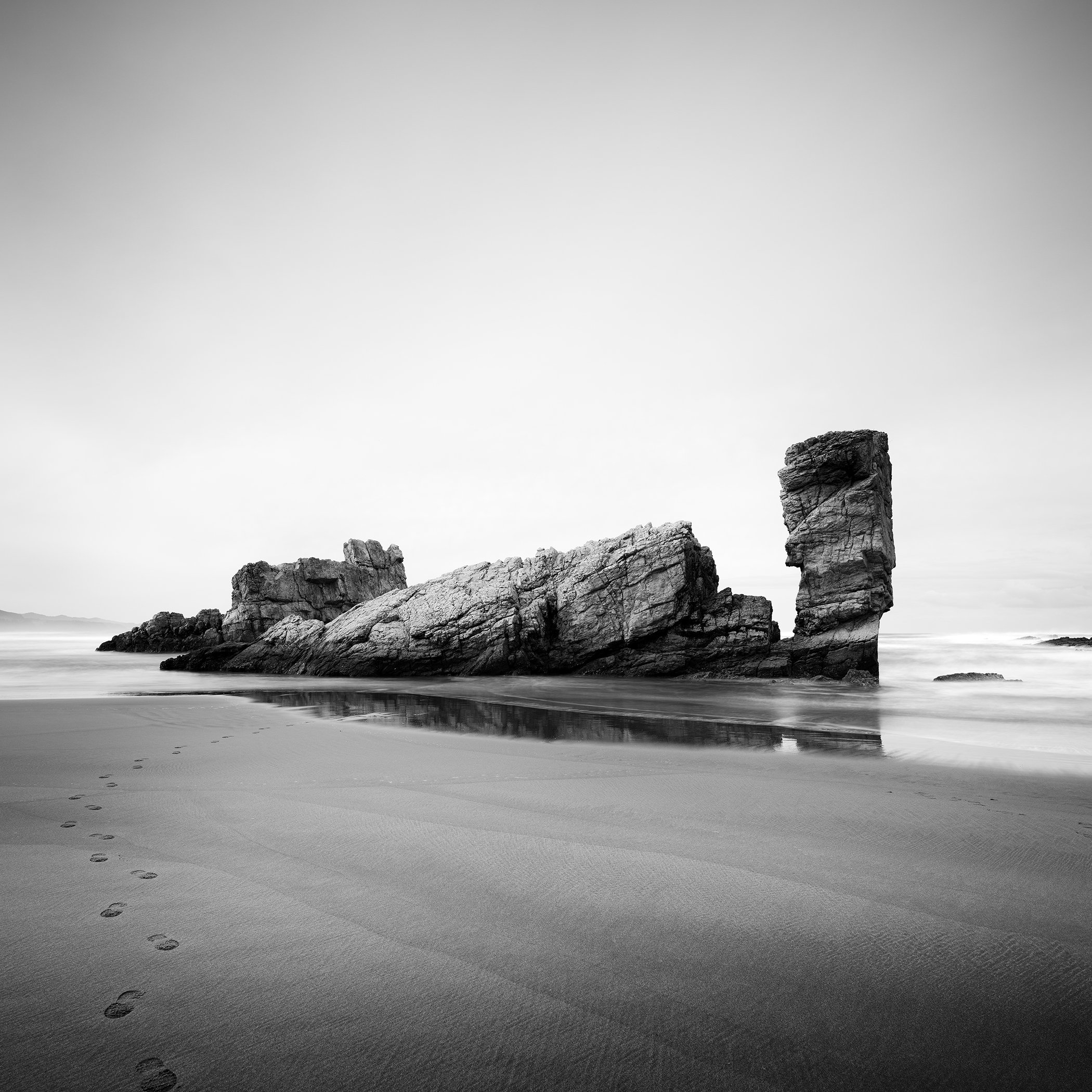 Bay of Biscay Study 4, minimalist black-and-white photograph of footprints, wet sand and a reflected sea stack in Spain by Gerald Berghammer.