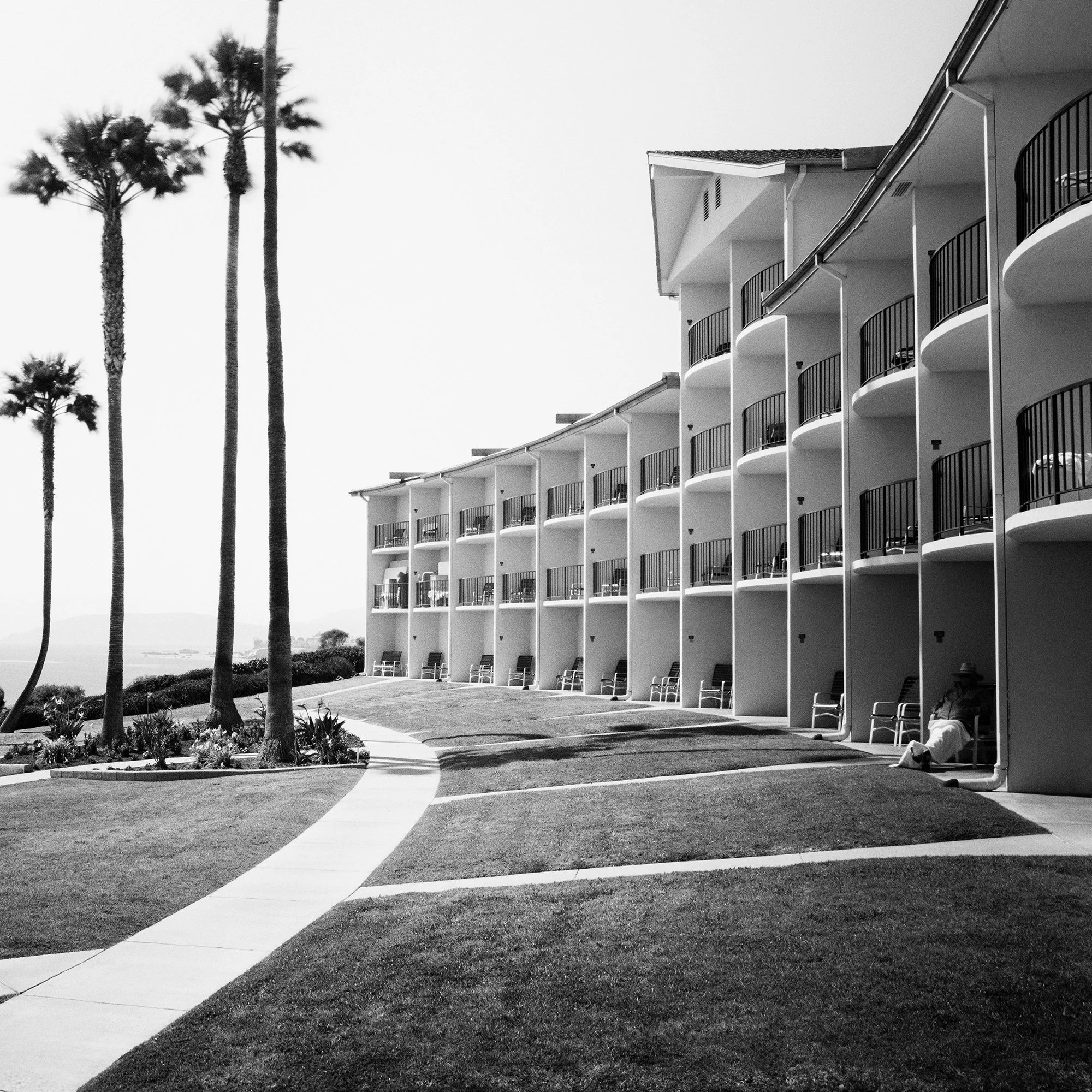 Gerald Berghammer - Black and white landscape photography. Seaside hotel with multiple balconies, a curved walkway, and tall palm trees near the ocean. Print detail 2