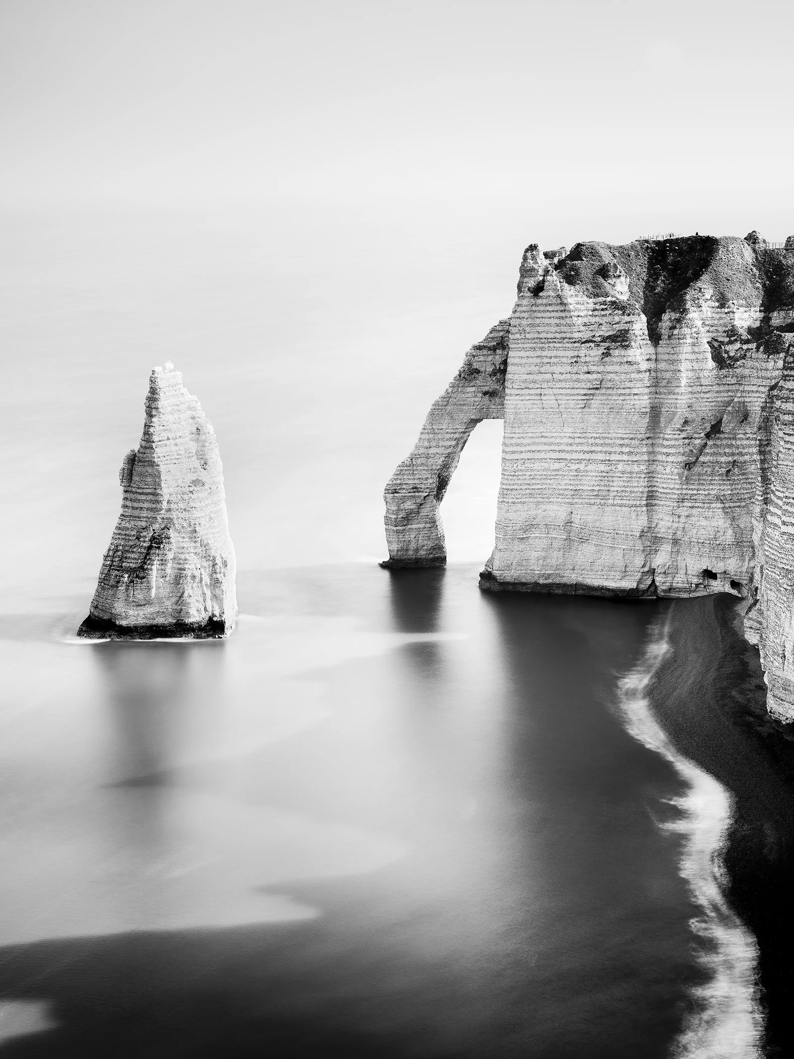 Black-and-white long-exposure photograph of the sea arch, chalk cliffs and Aiguille at Étretat in Normandy, France. Photo by Gerald Berghammer.
