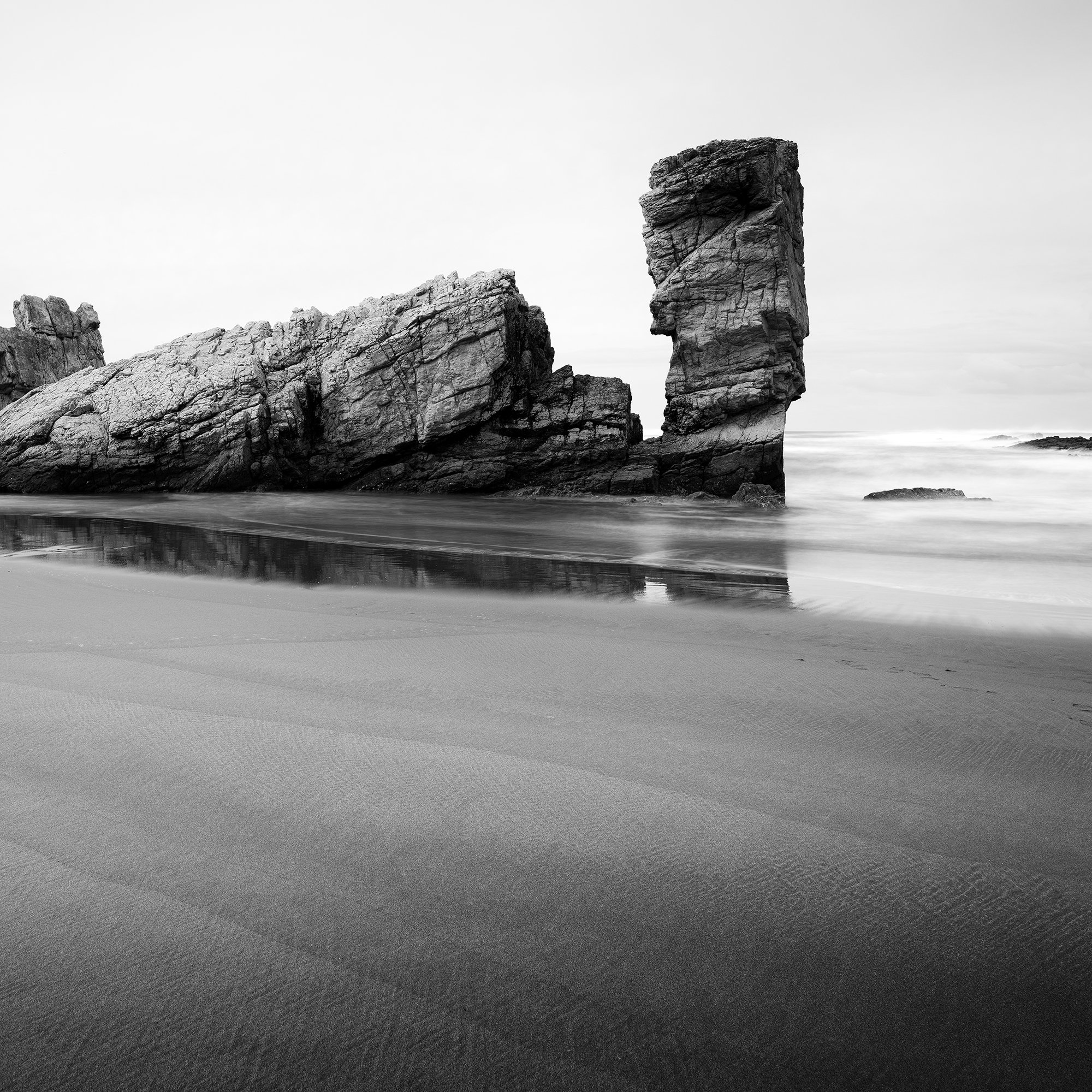© 2023 Gerald Berghammer. Black-and-white beach scene with wet sand, faint footprints, and a tall rock formation near the shore reflected in shallow water. Print detail 2