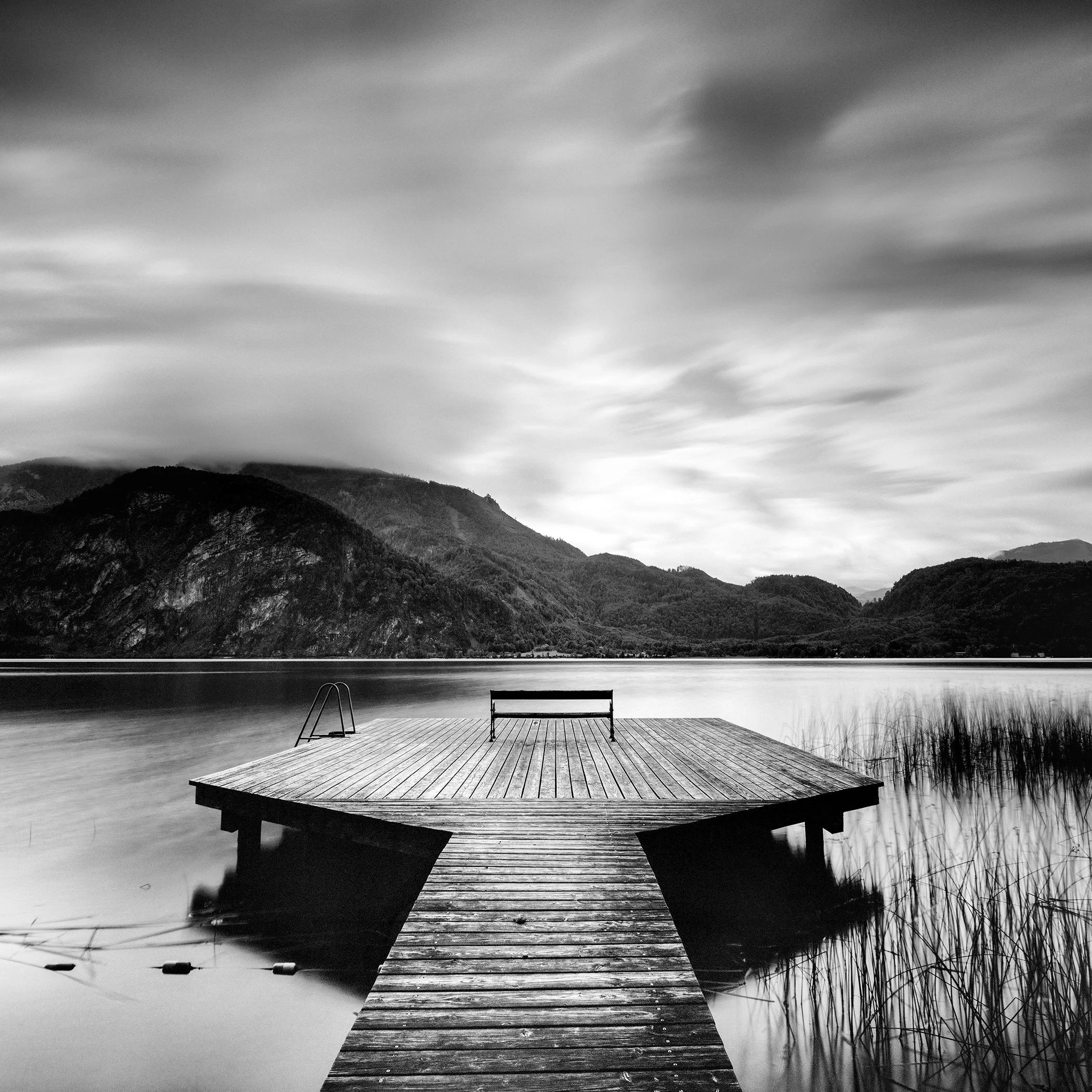 Black and white fine art photograph by Gerald Berghammer of a wooden pier on a calm lake with mountains and dramatic clouds.
