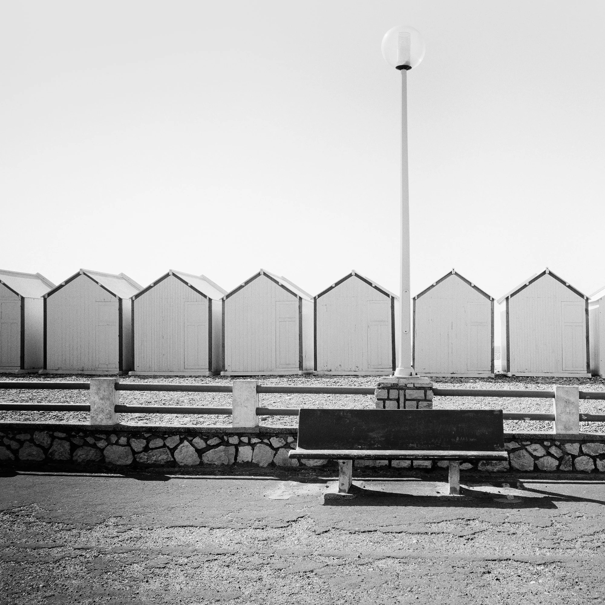 Detail of Bench on the Promenade Study 1 showing the empty bench, stone wall and left-hand beach huts in France by Gerald Berghammer.