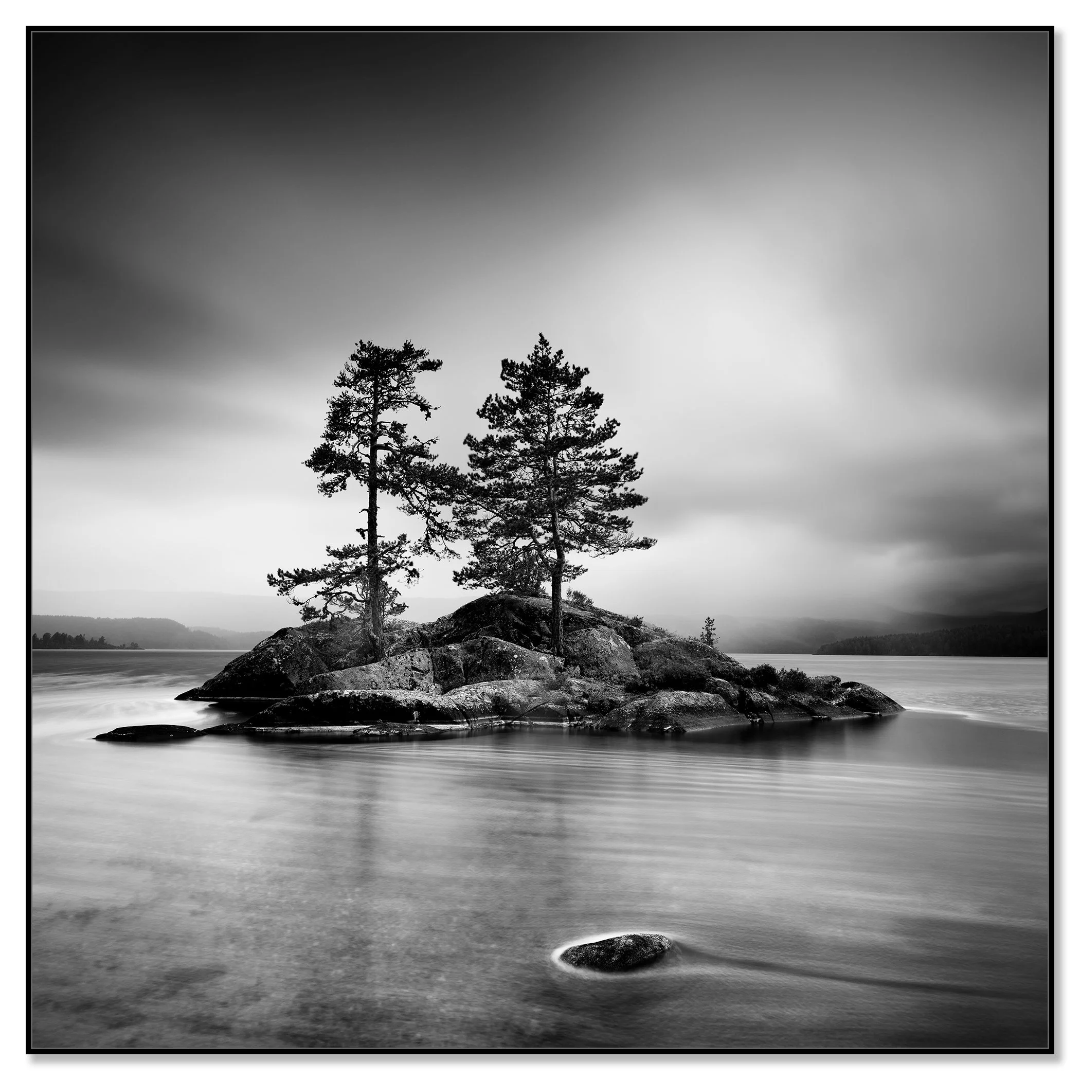 Black and white nature photograph of a rocky lake island with two pine trees and dramatic overcast skies – framed ArtBox black
