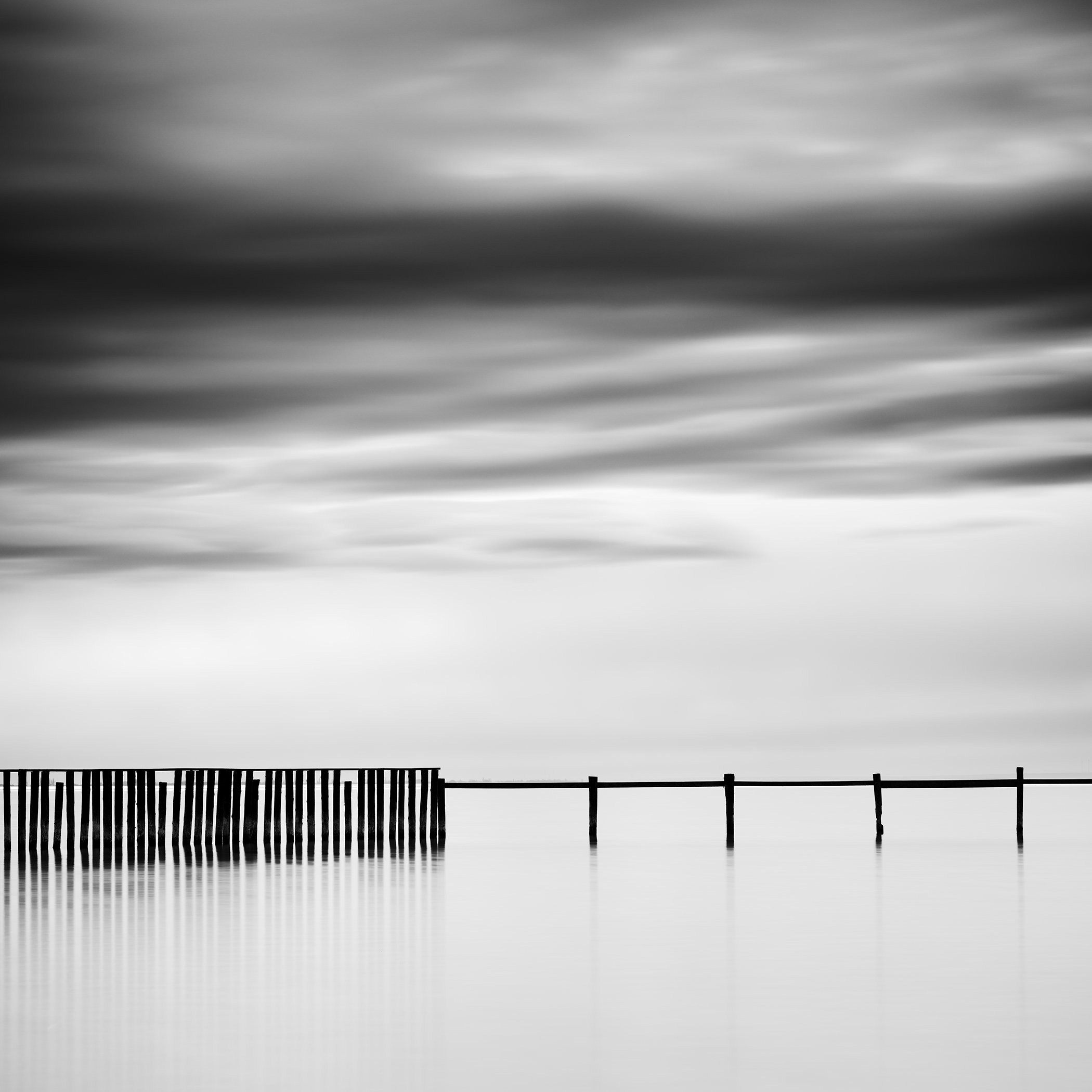 Minimalist black-and-white waterscape of a wooden swimming enclosure beneath blurred clouds on Lake Neusiedl, Austria.