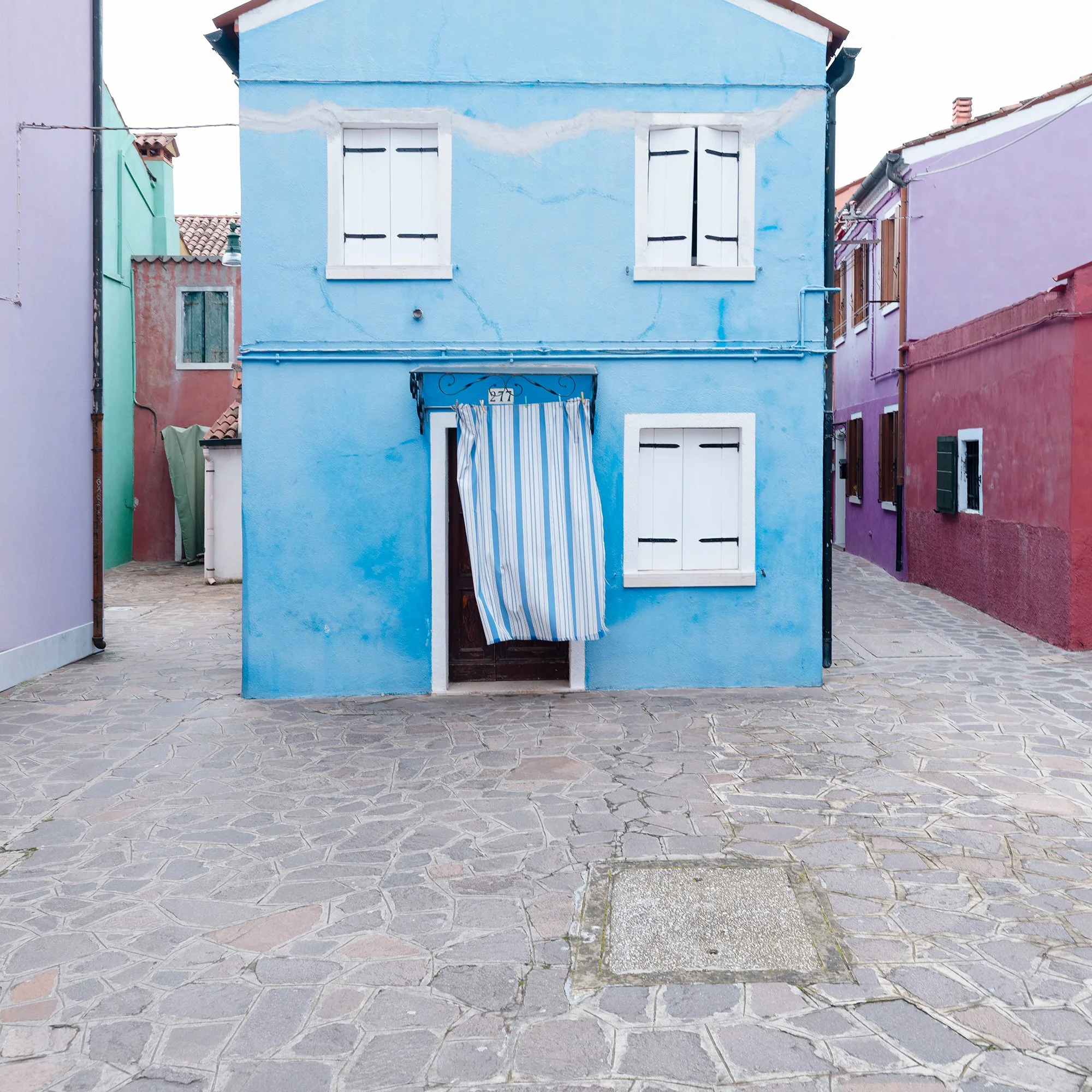 © 2025 Gerald Berghammer photo - Colorful houses in pastel shades on a cobblestone street, with colorful windows and a striped curtain hanging in front of one house. Print detail 1