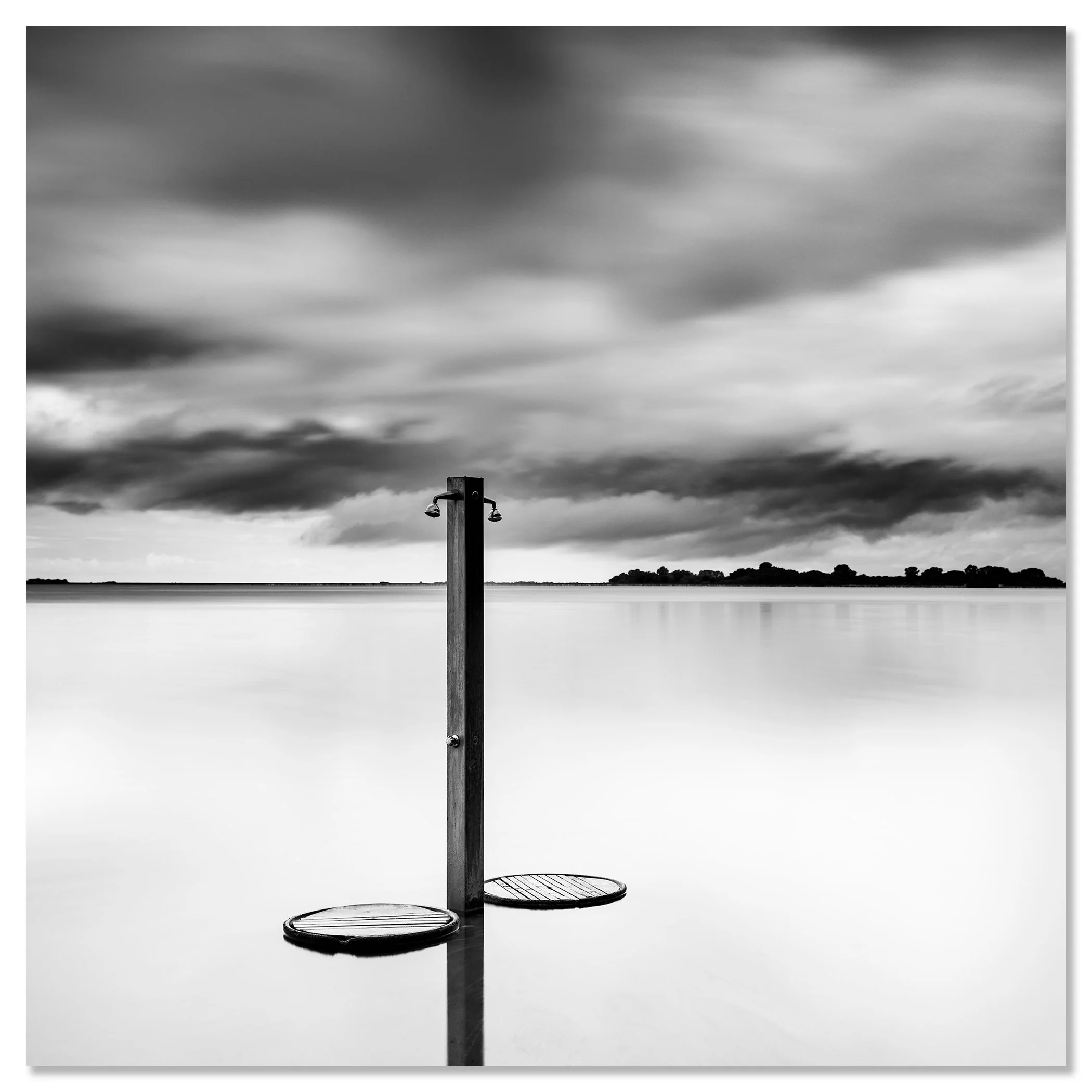 Black-and-white photo of a beach shower standing in calm water under dramatic storm clouds – dibond frameless