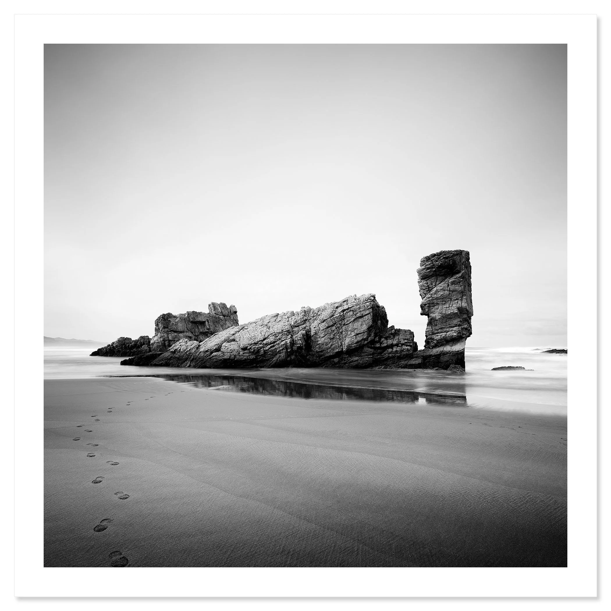 © 2023 Gerald Berghammer. Black-and-white beach scene with wet sand, faint footprints, and a tall rock formation near the shore reflected in shallow water. Fine art print only