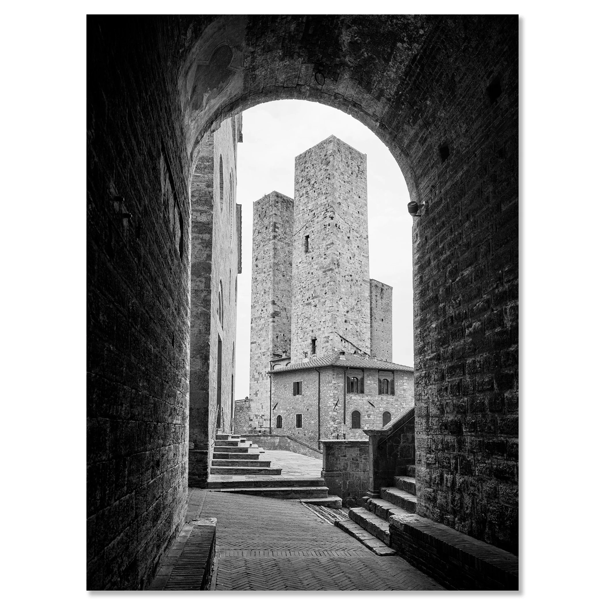 Black-and-white photograph of a medieval stone tower and buildings in San Gimignano, viewed through an arched stone gate – dibond frameless