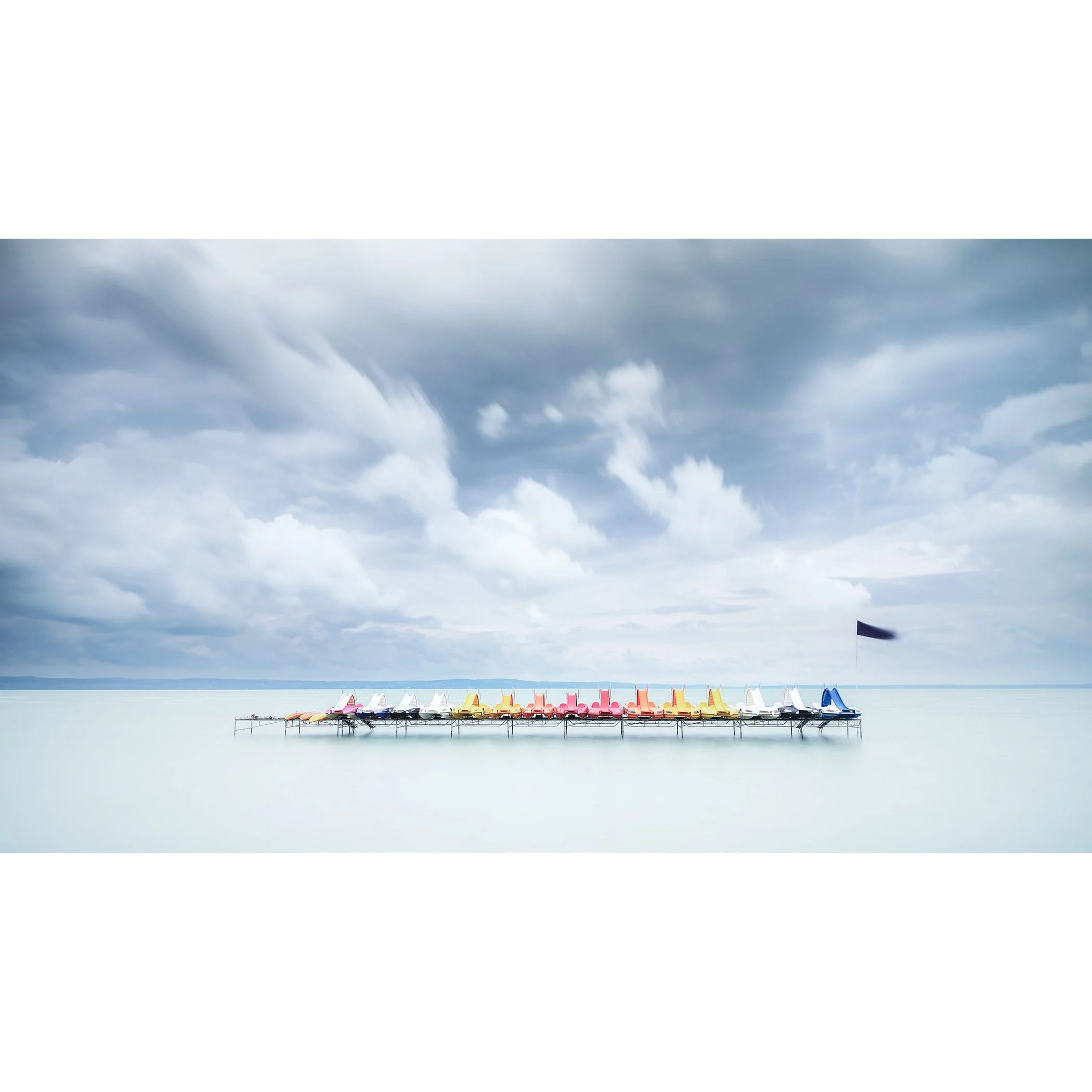 Gerald Berghammer - Color long exposure waterscape photography. Colorful paddle boats lined up on a dock over calm water, under a cloudy sky.