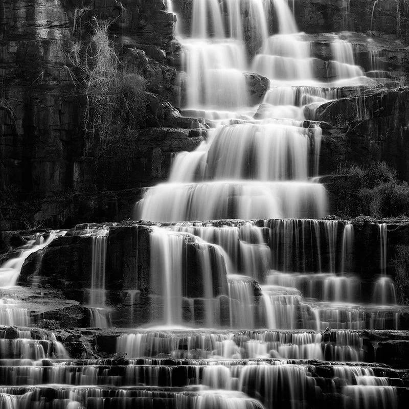 Black and white long-exposure photograph of a multi-tiered waterfall cascading over layered rock formations by Gerald Berghammer