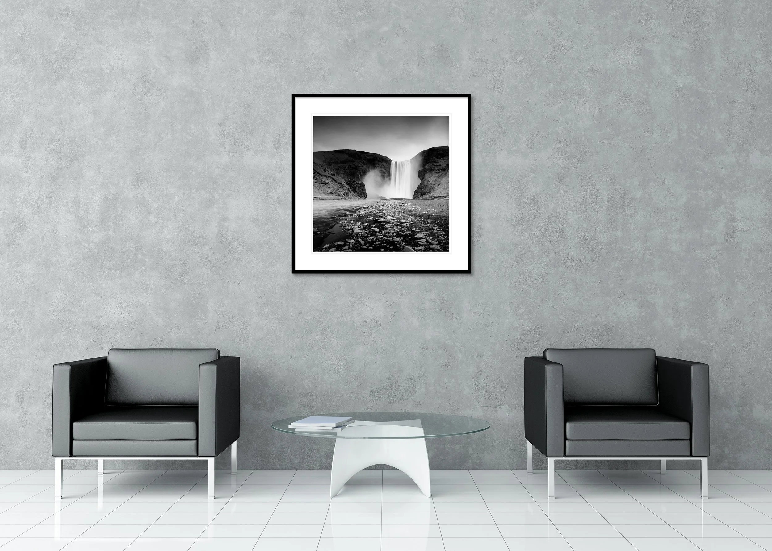 Black and white photograph of Skogafoss waterfall in Iceland cascading between dark volcanic cliffs with a rocky riverbed in the foreground, presented as an archival pigment print in a classic frame with passe-partout.