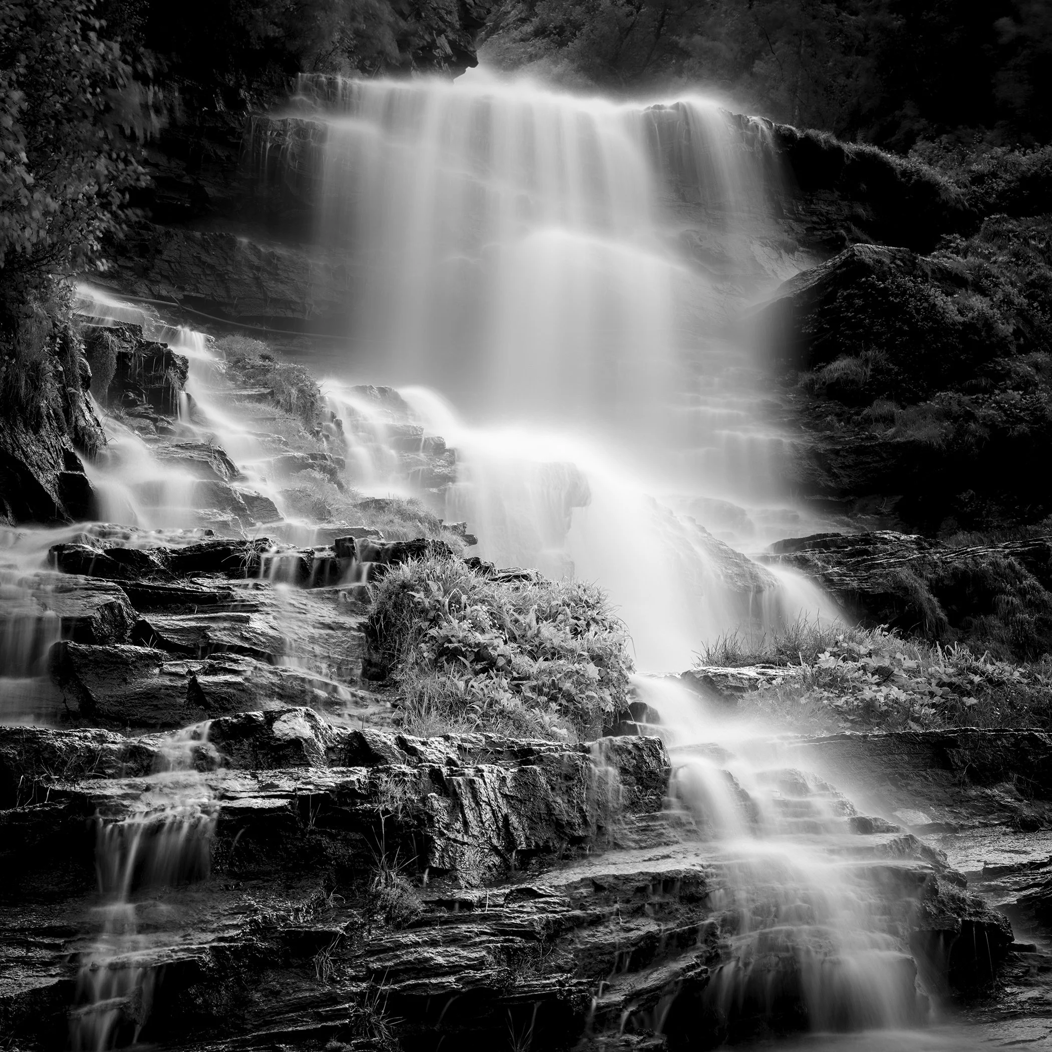 Black-and-white long-exposure waterfall over layered rocks, with mist-like water and dark textured stone.
