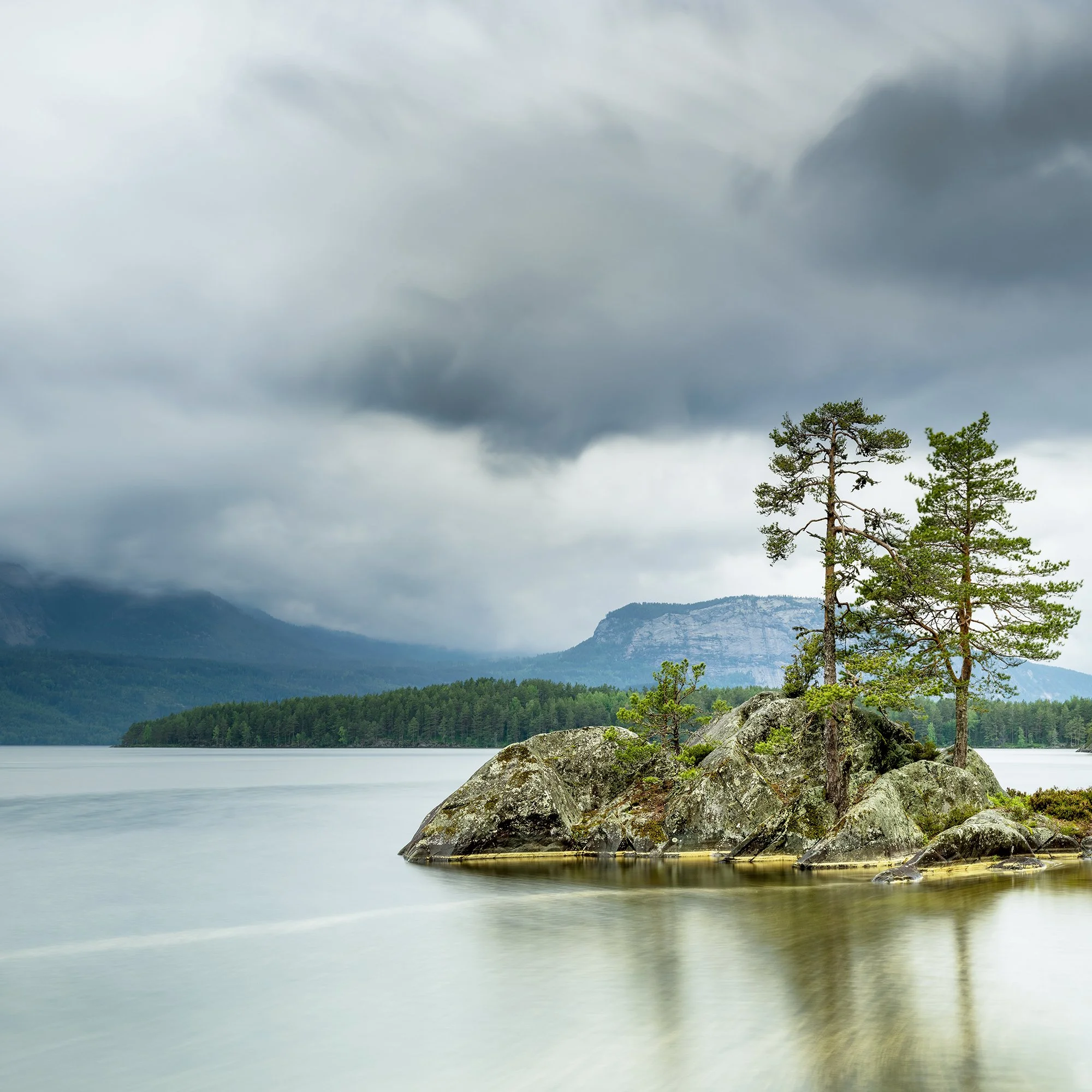 Gerald Berghammer - Color Photography. A small rocky island with tall pine trees surrounded by a calm lake under a cloudy sky and mountains behind. Print detail 1