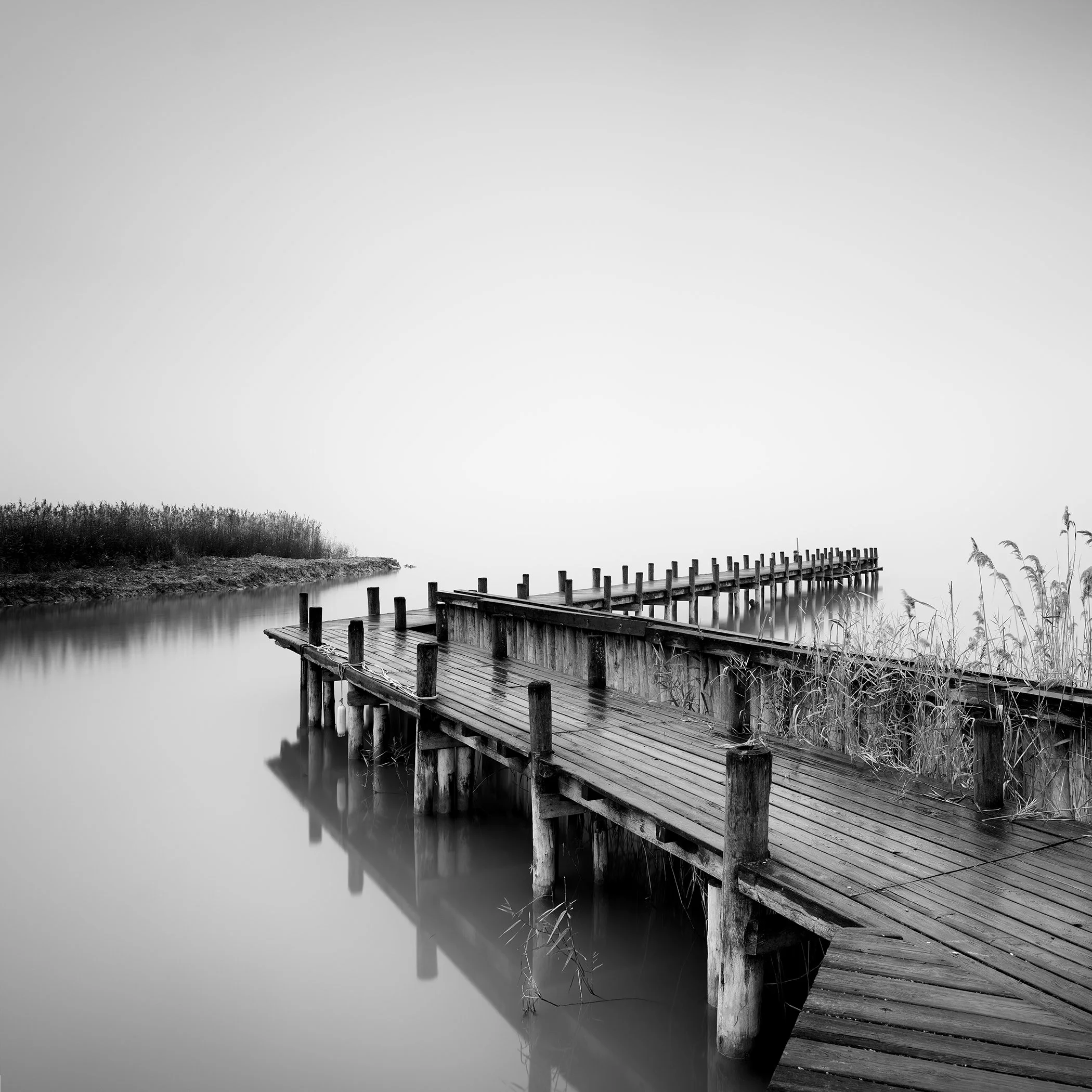 Black and white photograph of a wooden pier extending over calm lake water in a misty landscape