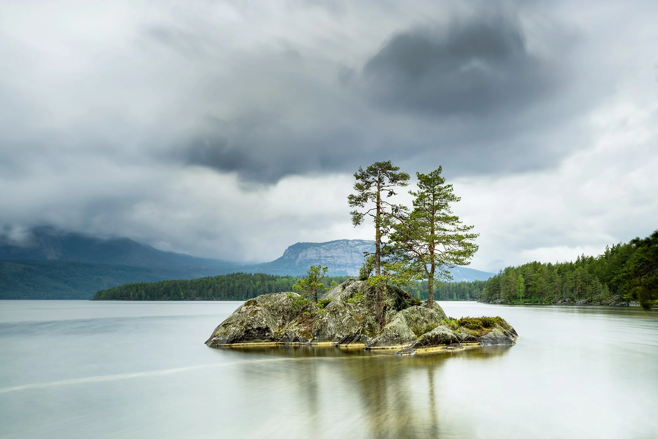 Small Rocky Island with Trees, Norway, long-exposure colour landscape with pine trees, calm lake and distant mountains.