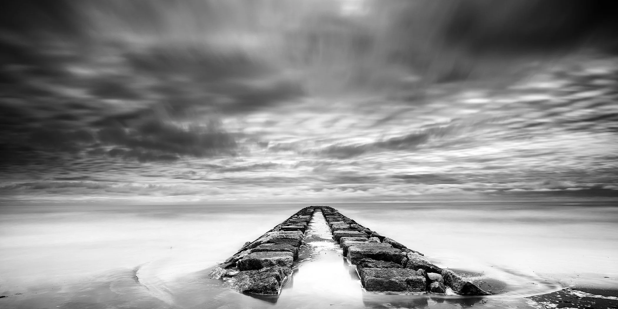 Black-and-white long-exposure panorama of a stone pier beneath dramatic clouds in Italy. Photo by Gerald Berghammer.