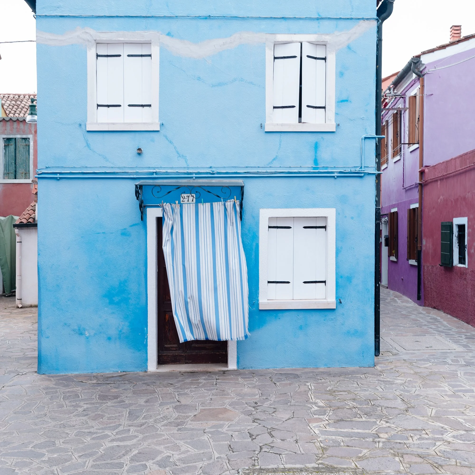 © 2025 Gerald Berghammer photo - Colorful houses in pastel shades on a cobblestone street, with colorful windows and a striped curtain hanging in front of one house. Print detail 3