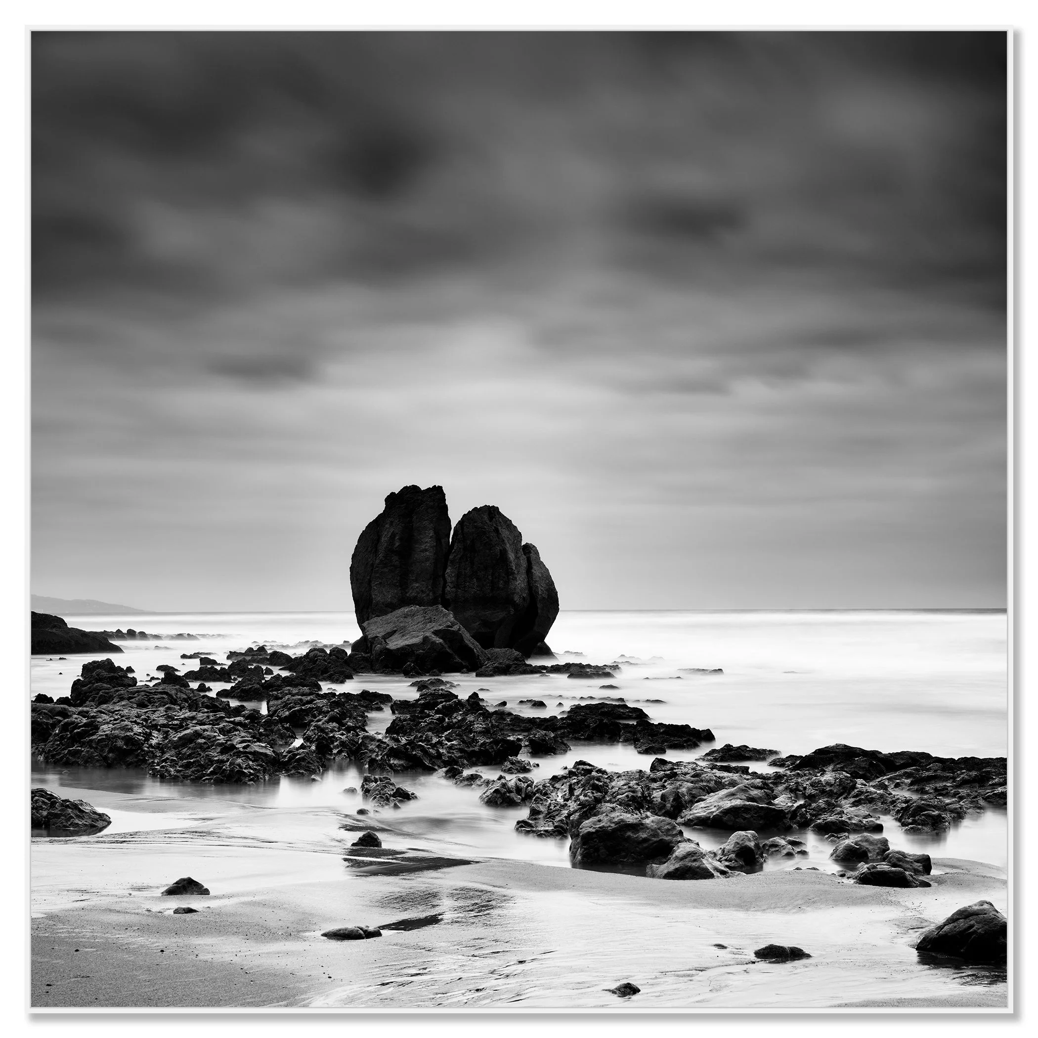 Black and white seascape with a large rock formation on a quiet beach under a cloudy sky – framed ArtBox white