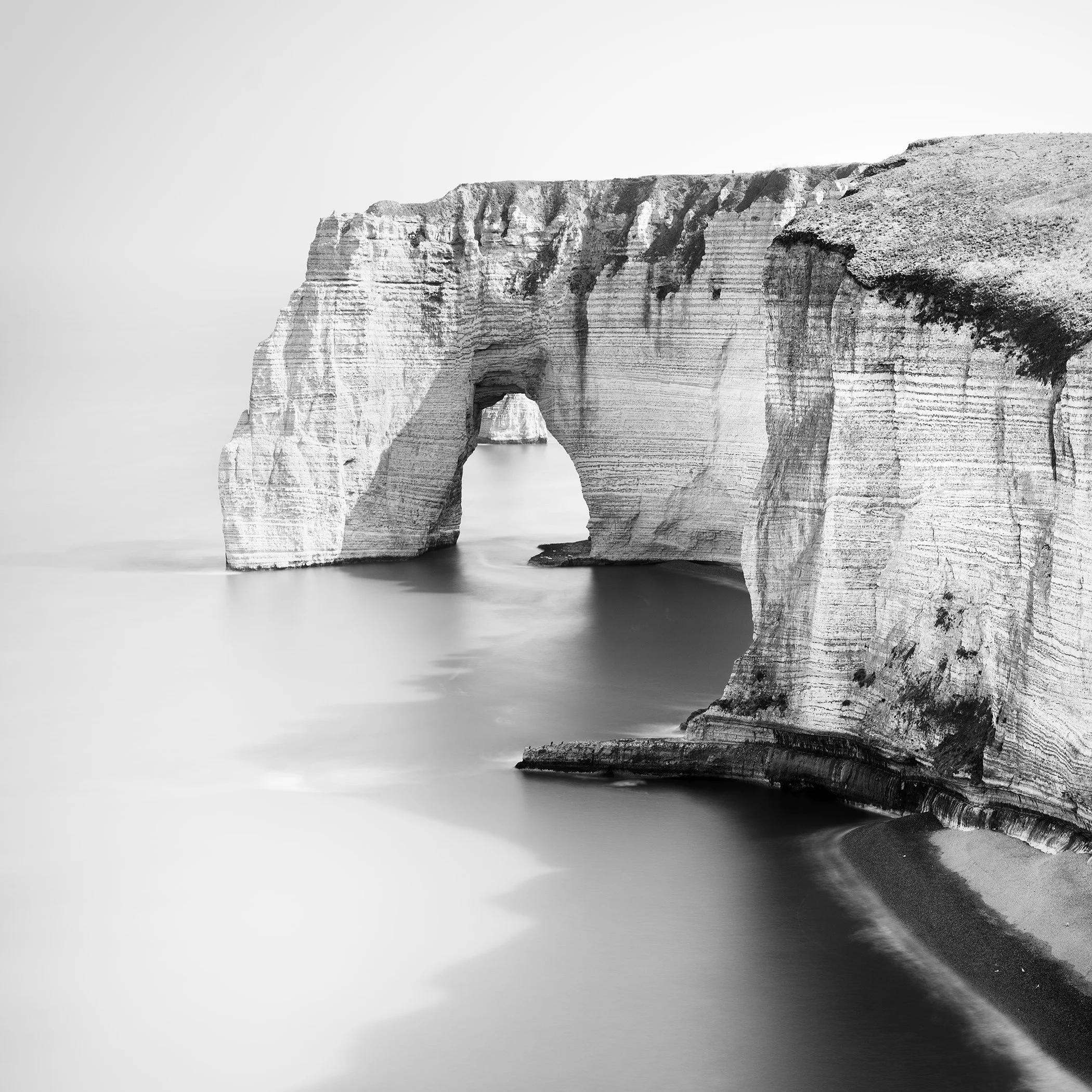 Black-and-white minimalist seascape of a natural rock arch along a rugged chalk coastline, with calm water blurred by long exposure.