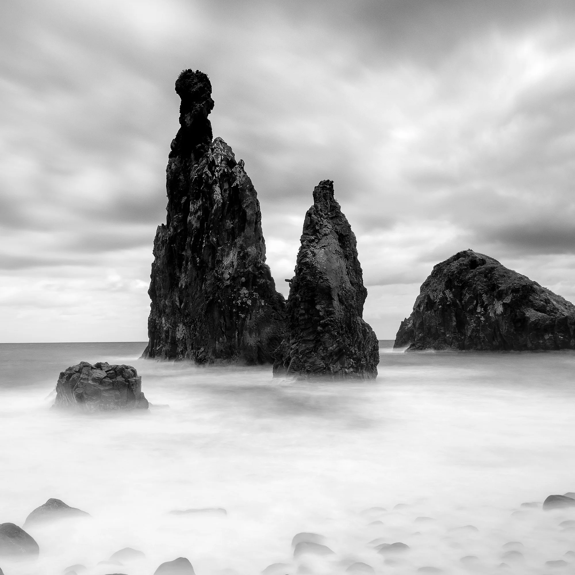 © 2021 Gerald Berghammer - Black and white seascape photography. Rocky seashore with three prominent sea stacks rising from the water, cloudy sky overhead. Print detail 3