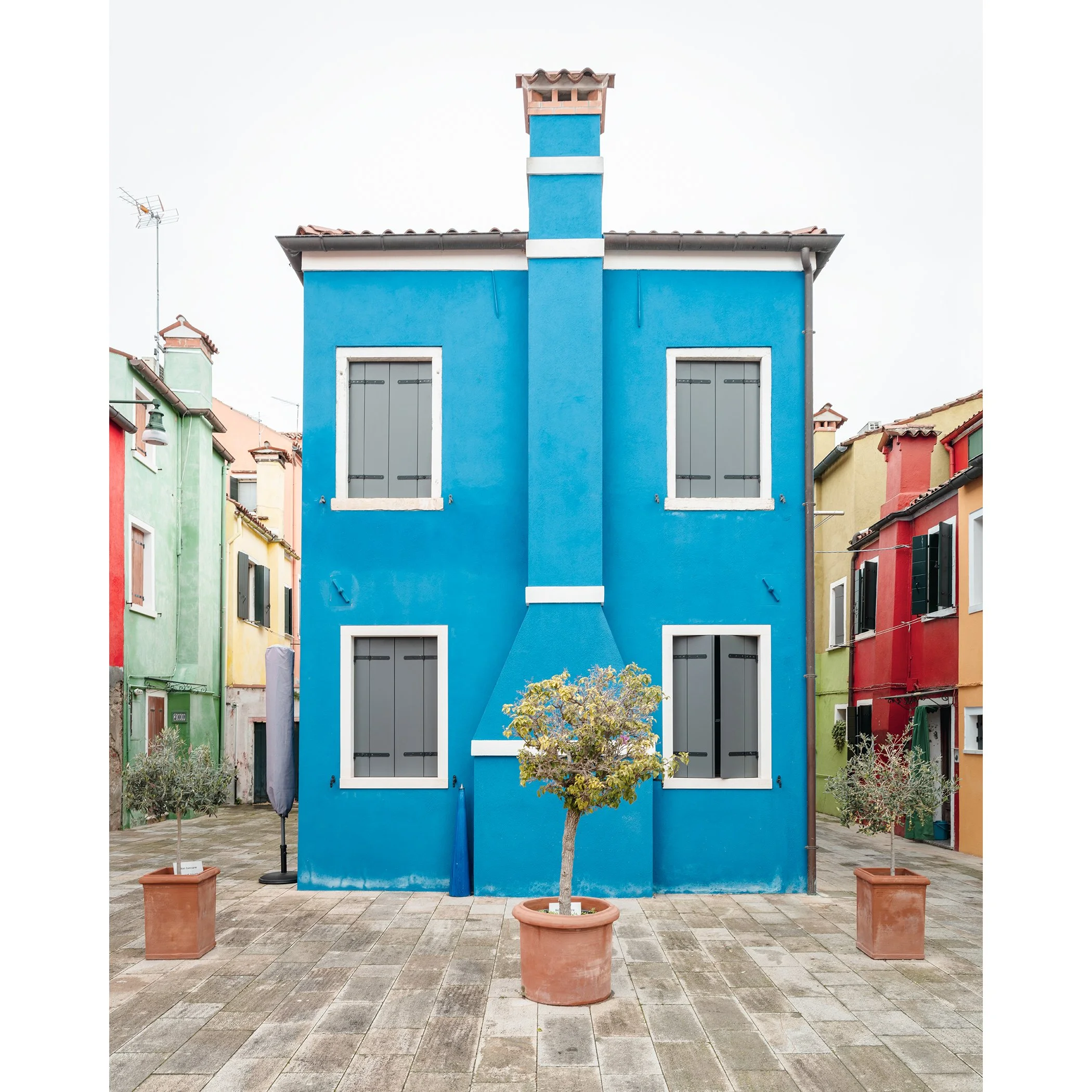 © 2025 Gerald Berghammer - Color cityscape photo. Colorful row houses with a prominent blue house in the center, potted small trees in front, and a paved courtyard.