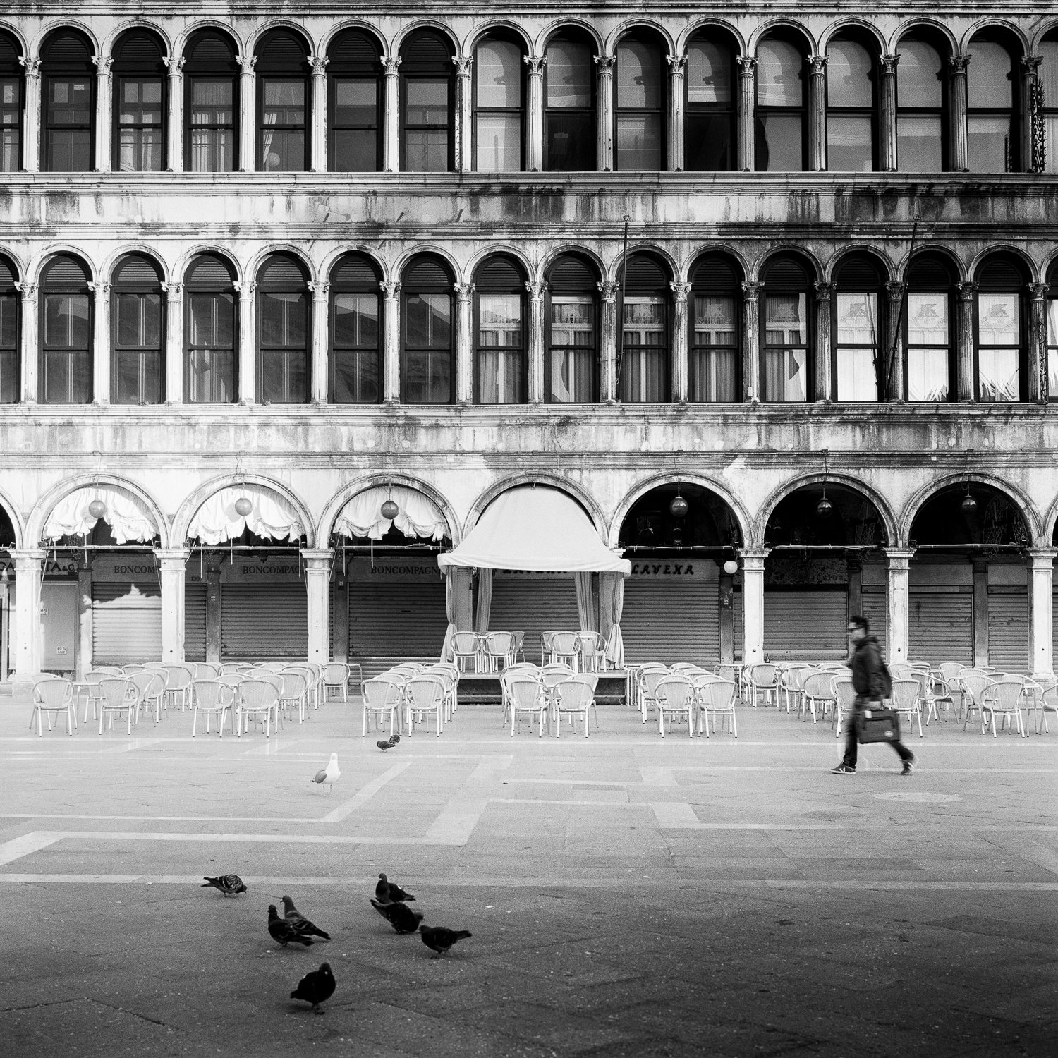 Black-and-white photograph of Caffè Florian in Piazza San Marco, Venice, with historic arcades, pigeons and a passer-by, by Gerald Berghammer.