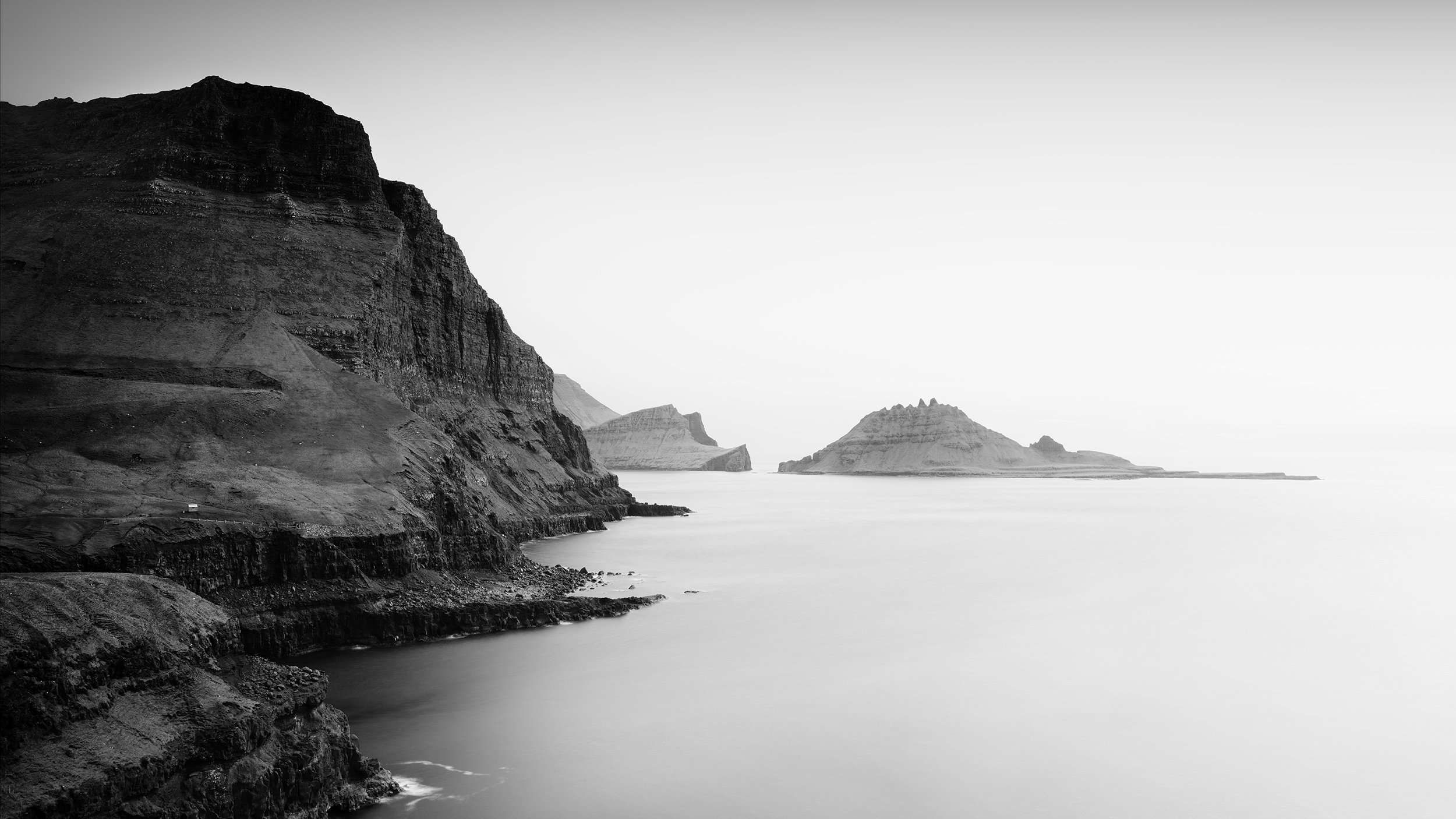 Monochrome seascape showing rugged cliff coastline and distant rocky islets under a bright sky
