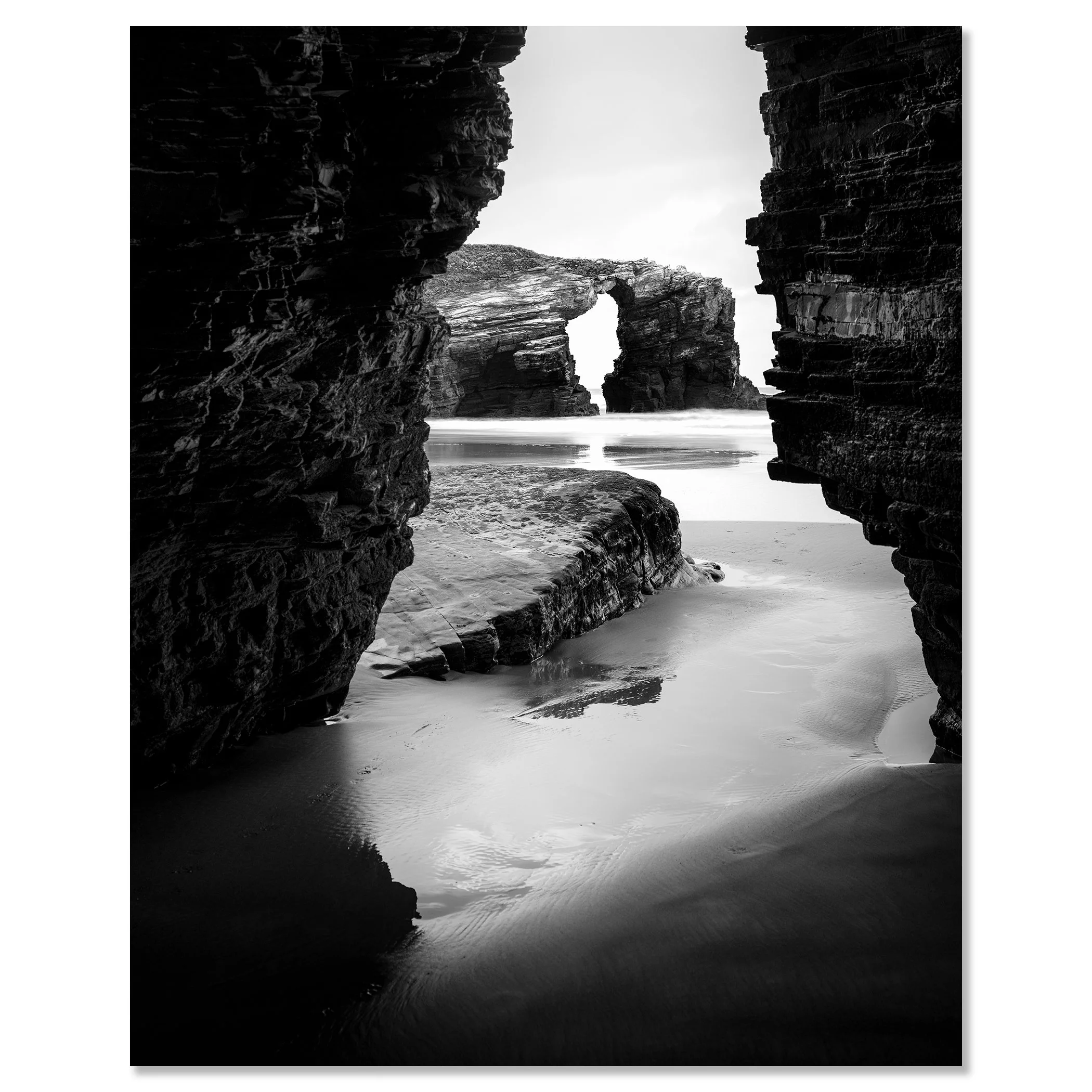Monochrome seascape photo of Catedrais Beach, Spain, showing a natural rock arch and ocean waves viewed through a narrow opening between large rocks – dibond frameless