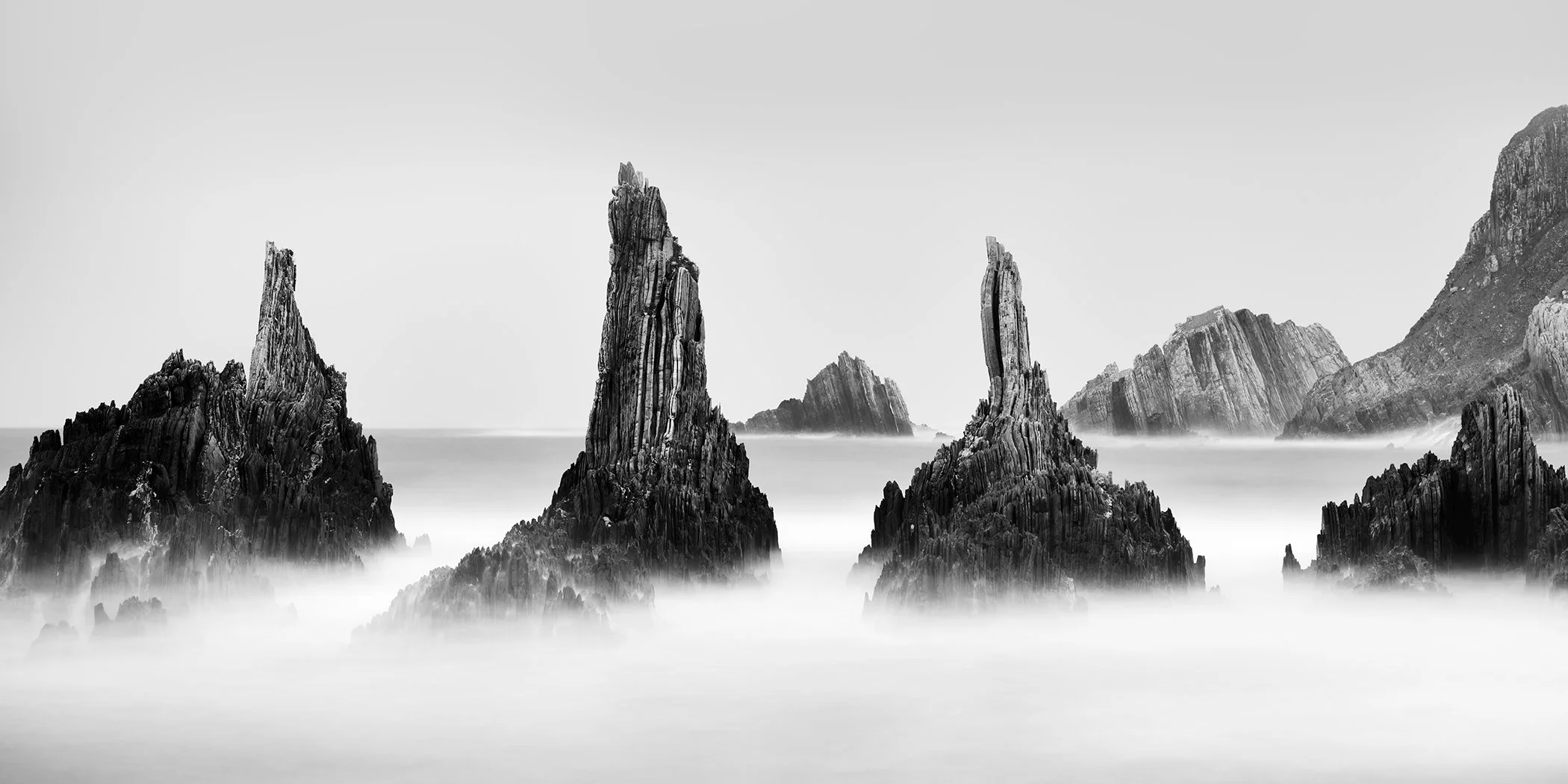 Monochrome coastal landscape showing dramatic rock formations, soft sea mist and distant cliffs in Spain by Gerald Berghammer.