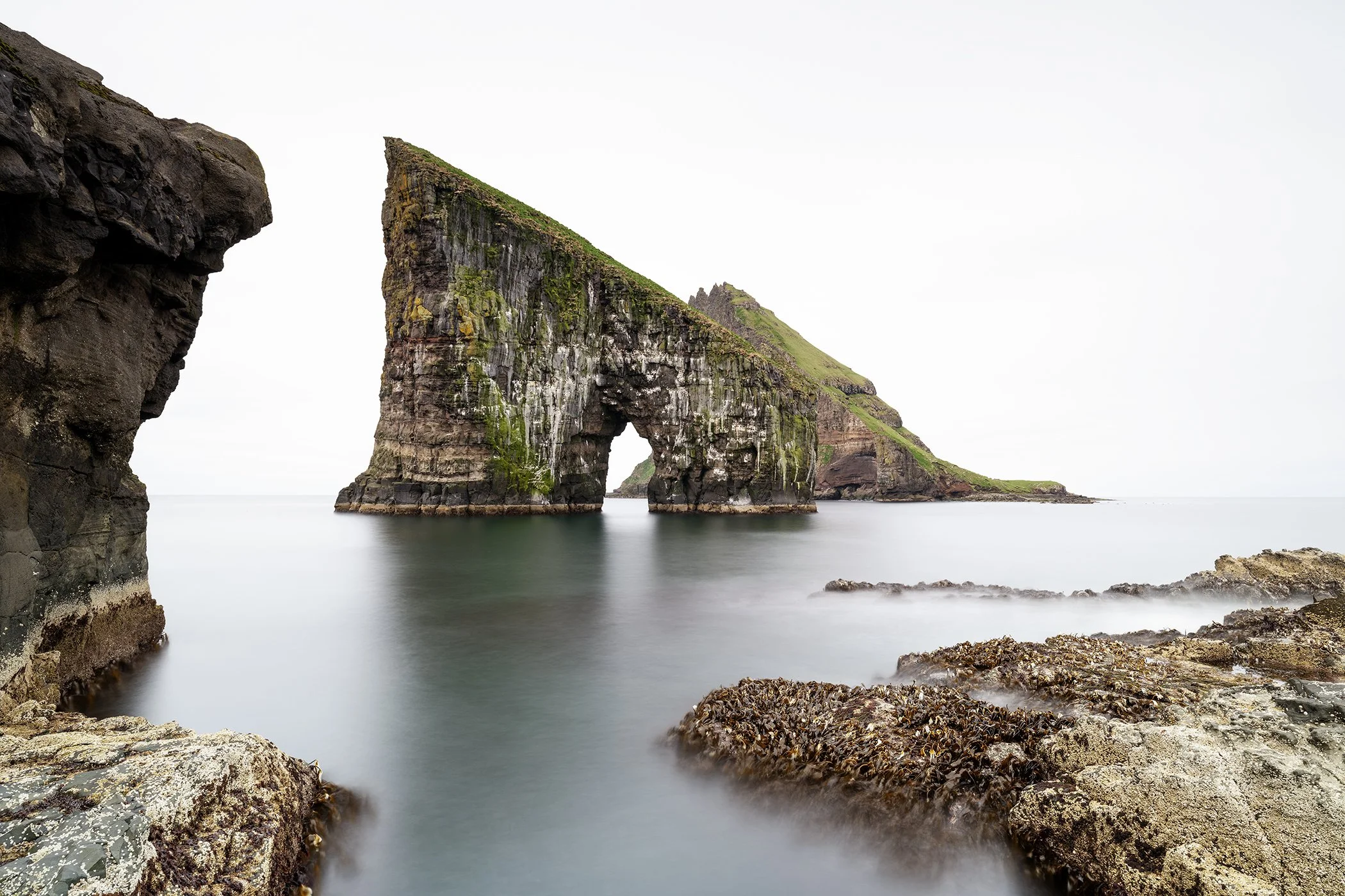 Colour fine art photograph of Drangarnir Sea Arch rising above calm North Atlantic water in the Faroe Islands by Gerald Berghammer.