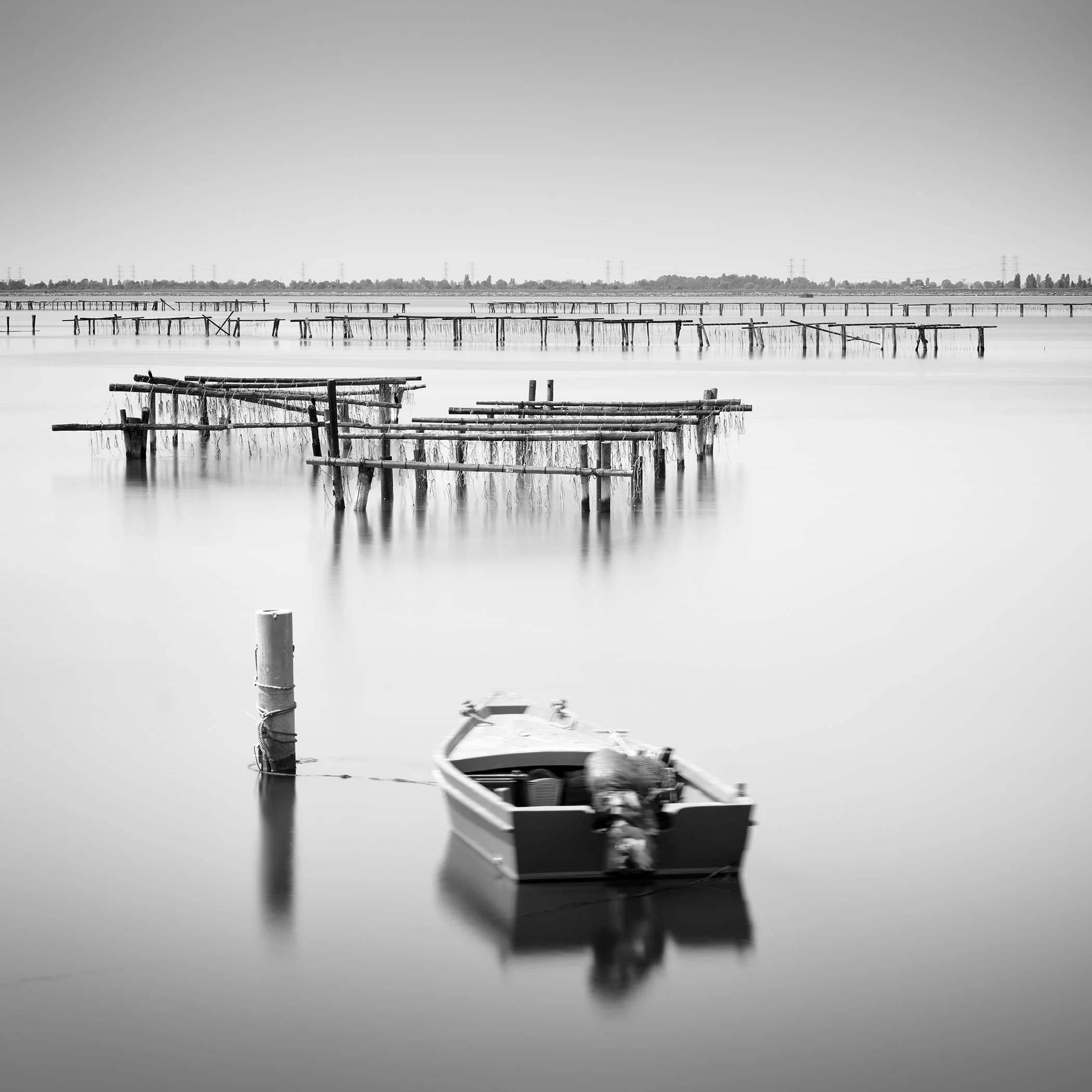 Gerald Berghammer - Black and White Seascape Photography. Small boat tied to a post on calm water, with wooden piers from an Aquaculture in Italy.
