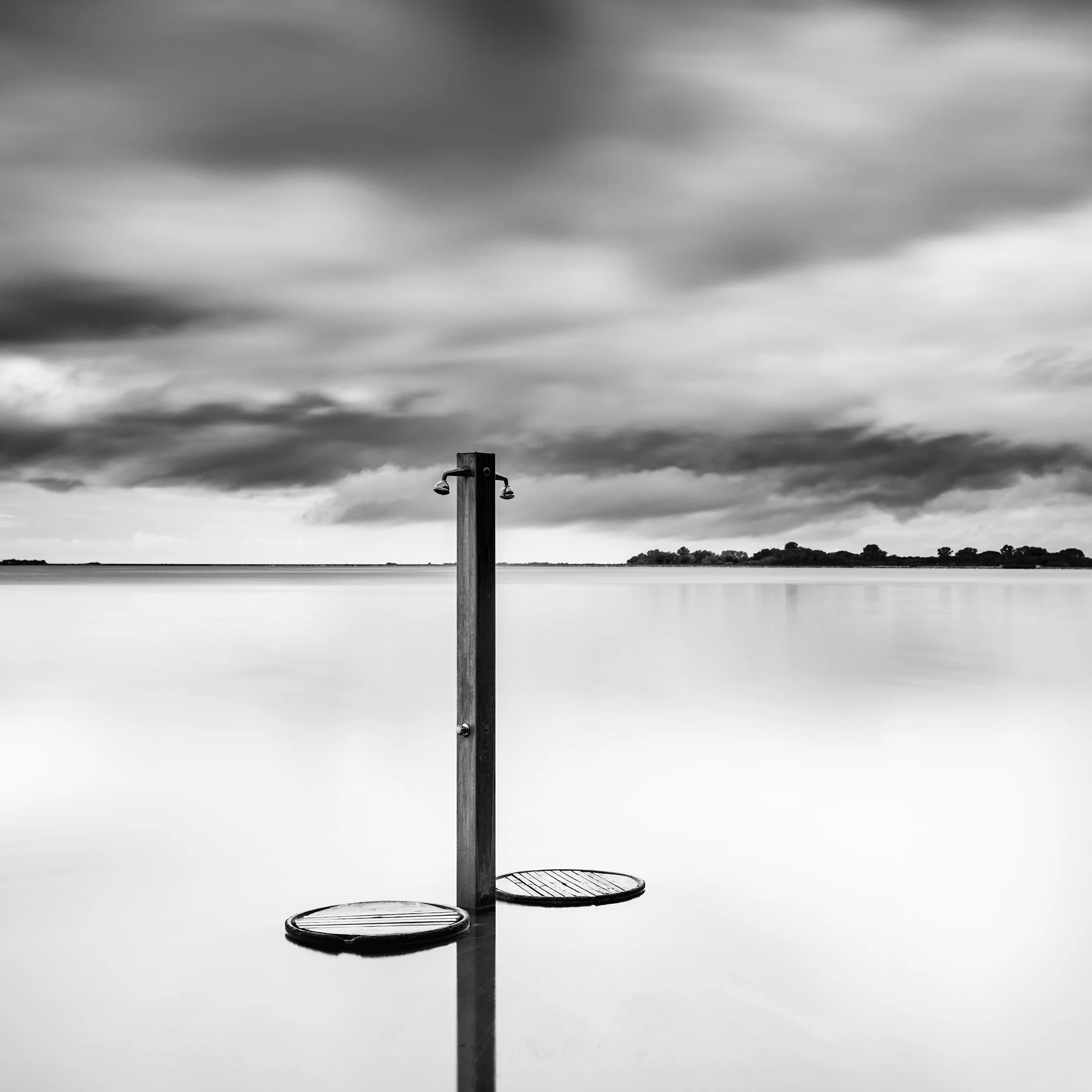 Beach Shower in a Storm – minimalist black-and-white seascape photograph of a solitary beach shower in still water beneath storm clouds in Tuscany, Italy, by Gerald Berghammer.