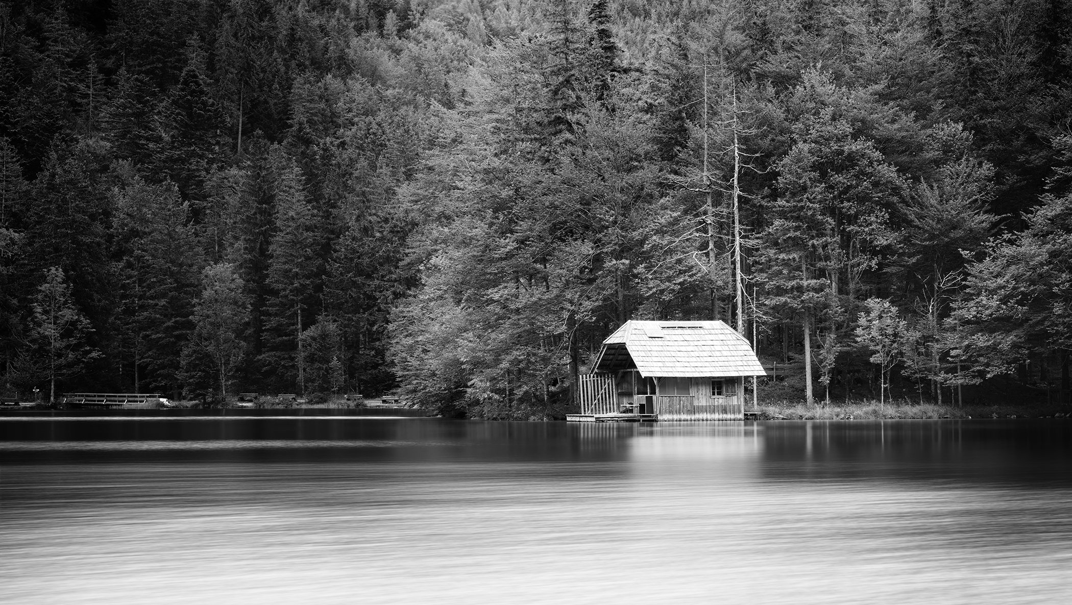 Black-and-white long-exposure photo of a quiet lake with mirror-smooth water and a boathouse at the forest edge.