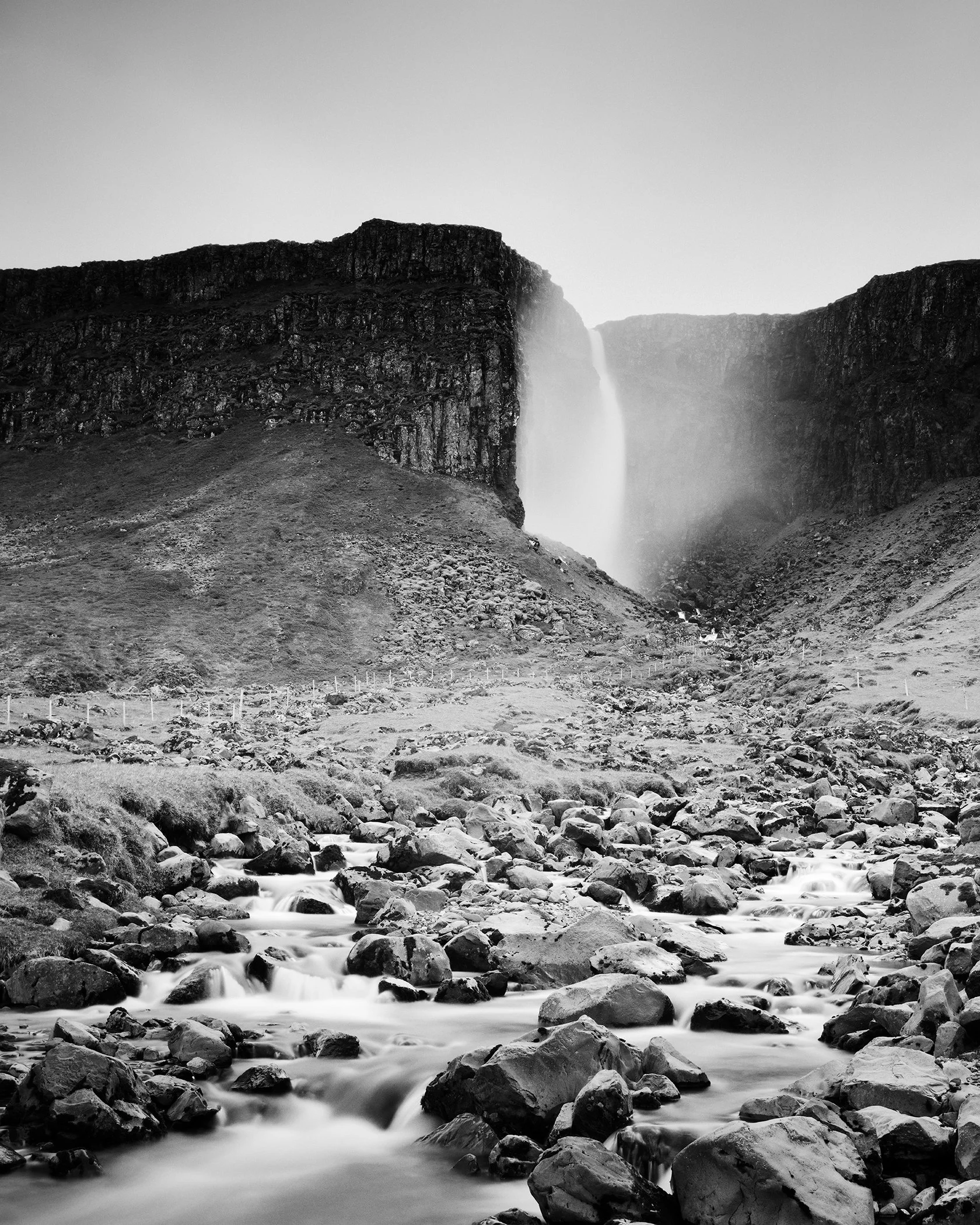 Black-and-white fine art photograph of an Icelandic waterfall fading into mist above a rugged rocky riverbed. Photo by Gerald Berghammer.