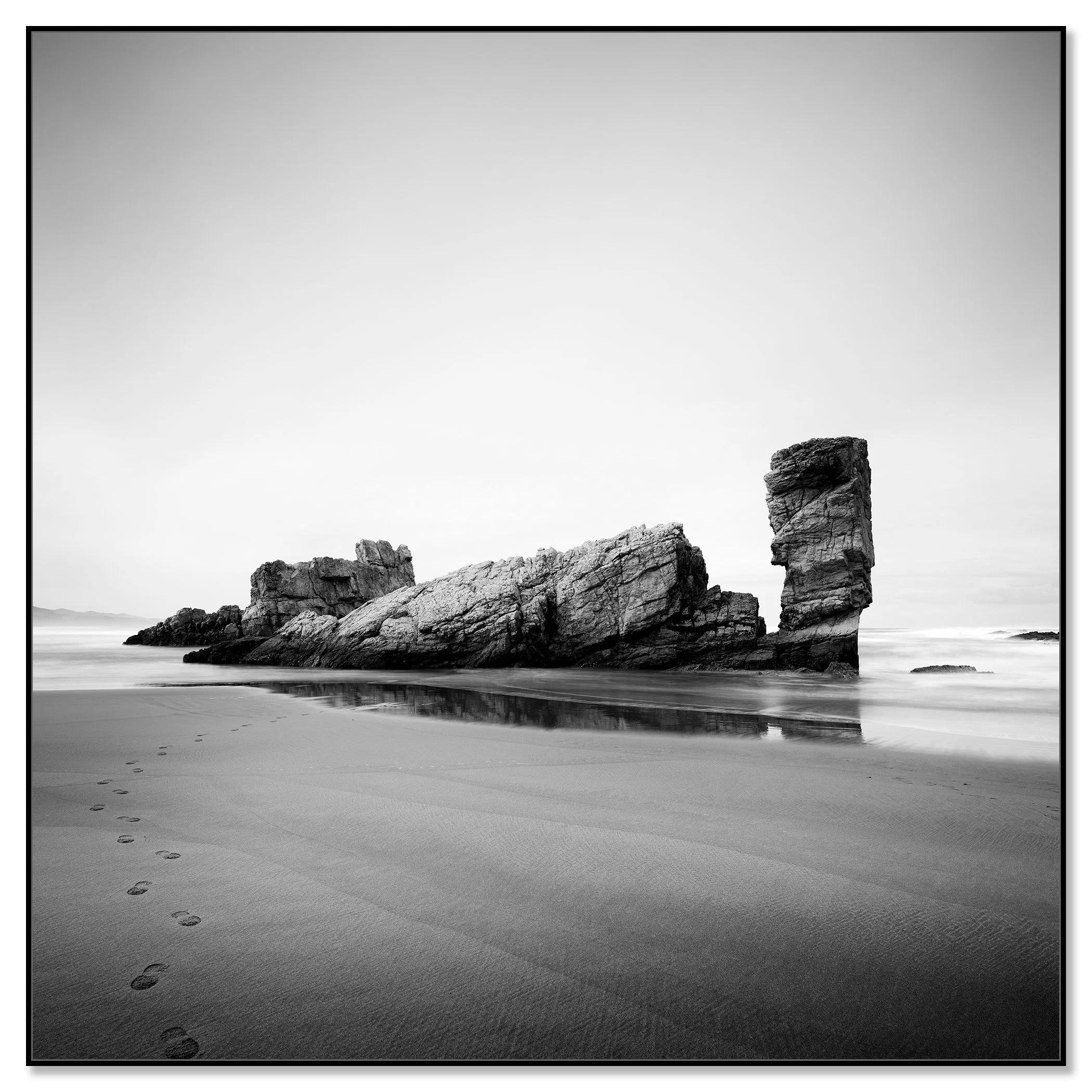 Black-and-white beach photograph with wet sand, faint footprints and a tall rock formation reflected in shallow water – framed ArtBox black